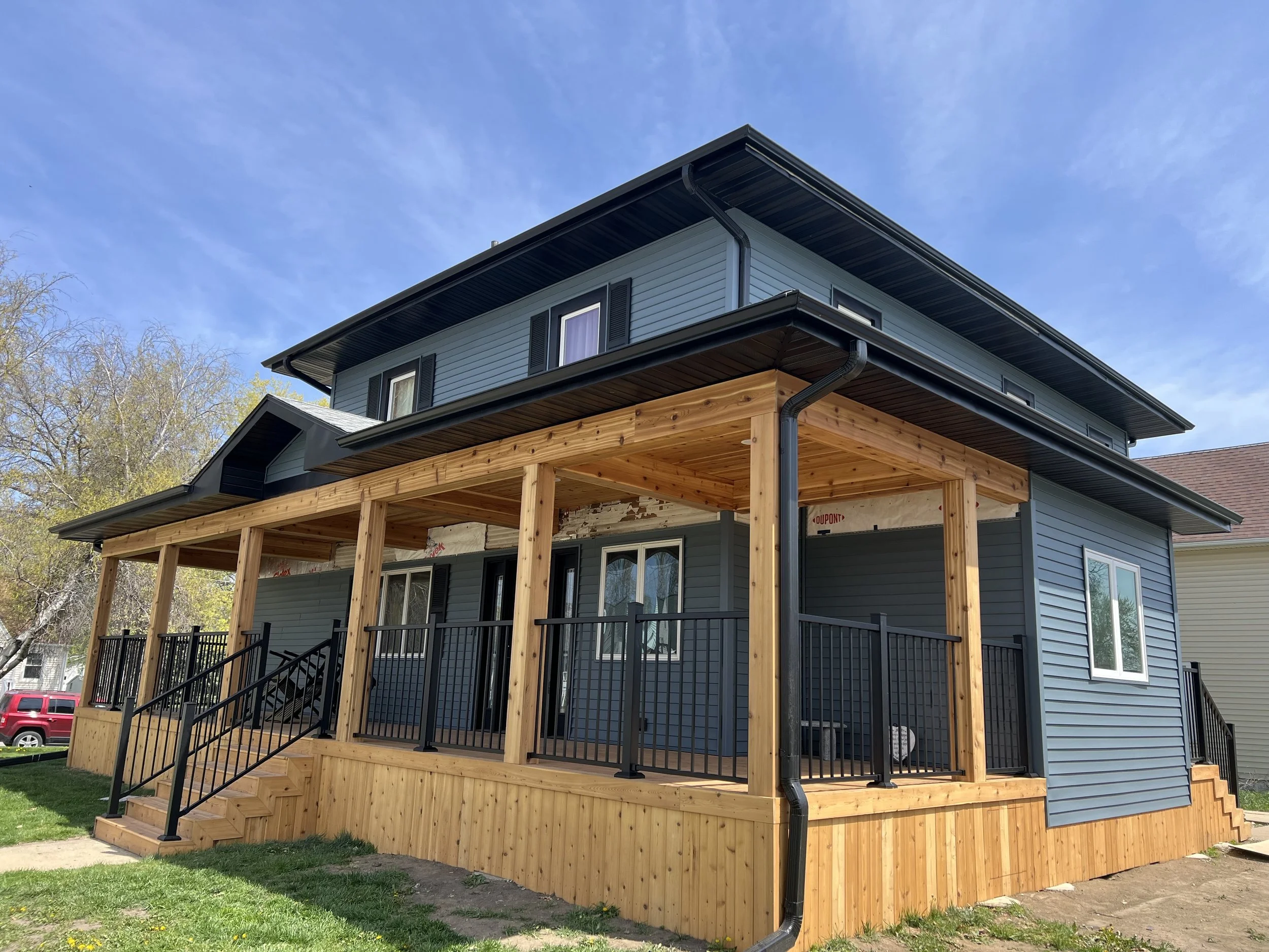 Back view of a blue house with three windows, a door, and a small wooden staircase leading to the door. The house has white gutters and trim, with a grassy lawn in the foreground and a clear blue sky above.