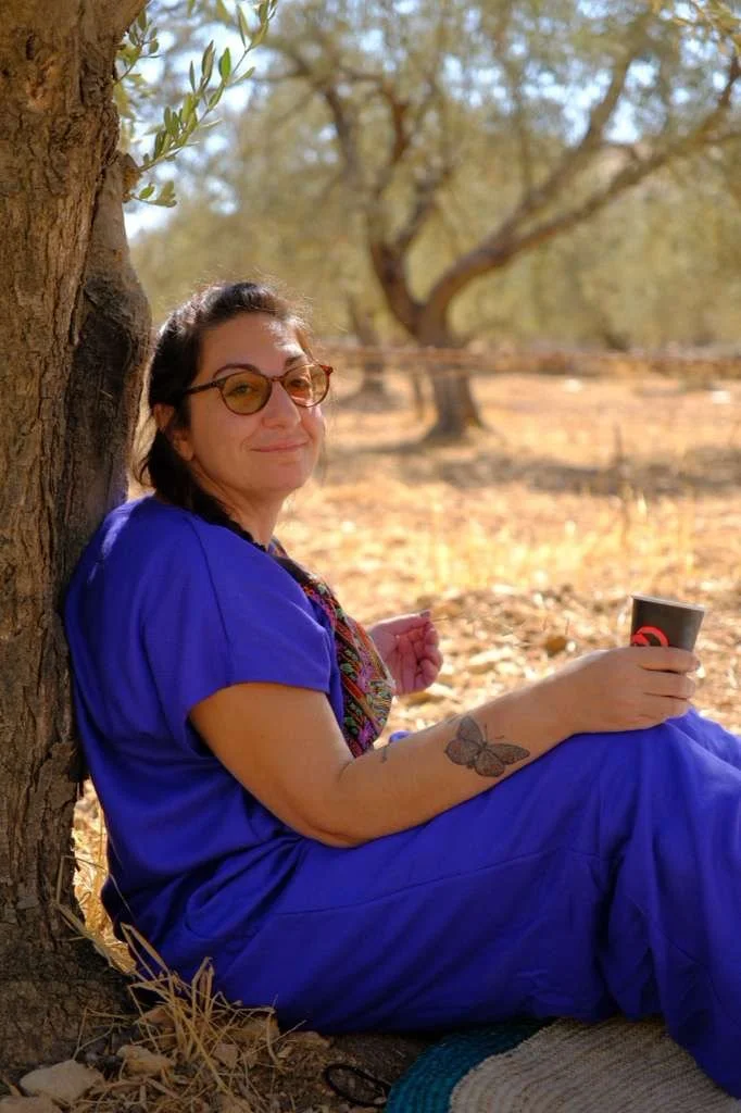 A woman in a blue dress with glasses sitting against a tree outdoors, holding a cup, with sunlight filtering through the trees.