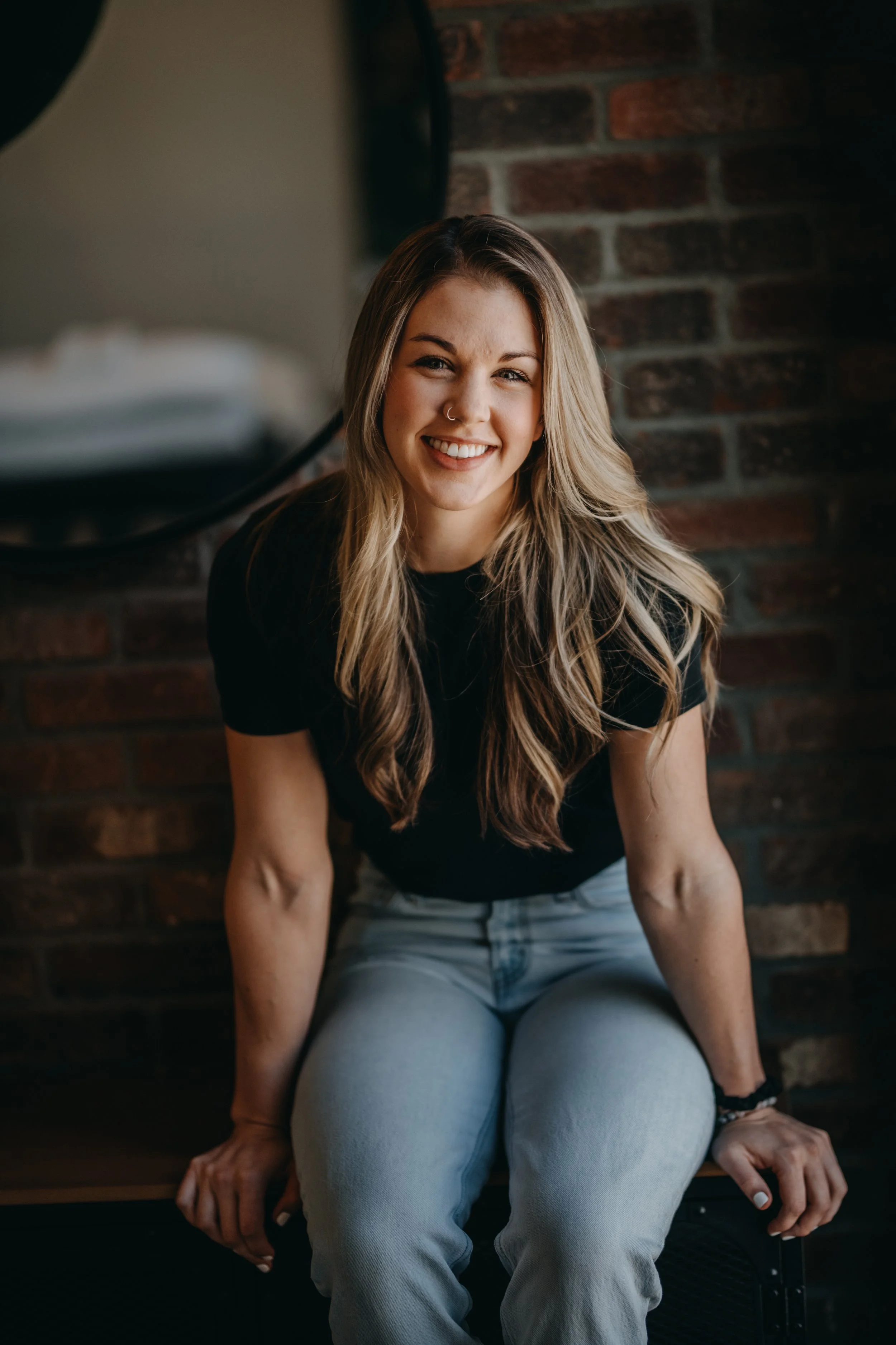 A young woman with long blonde hair, wearing a nose ring and a black top, sitting on a black chair in front of a brick wall, smiling at the camera.