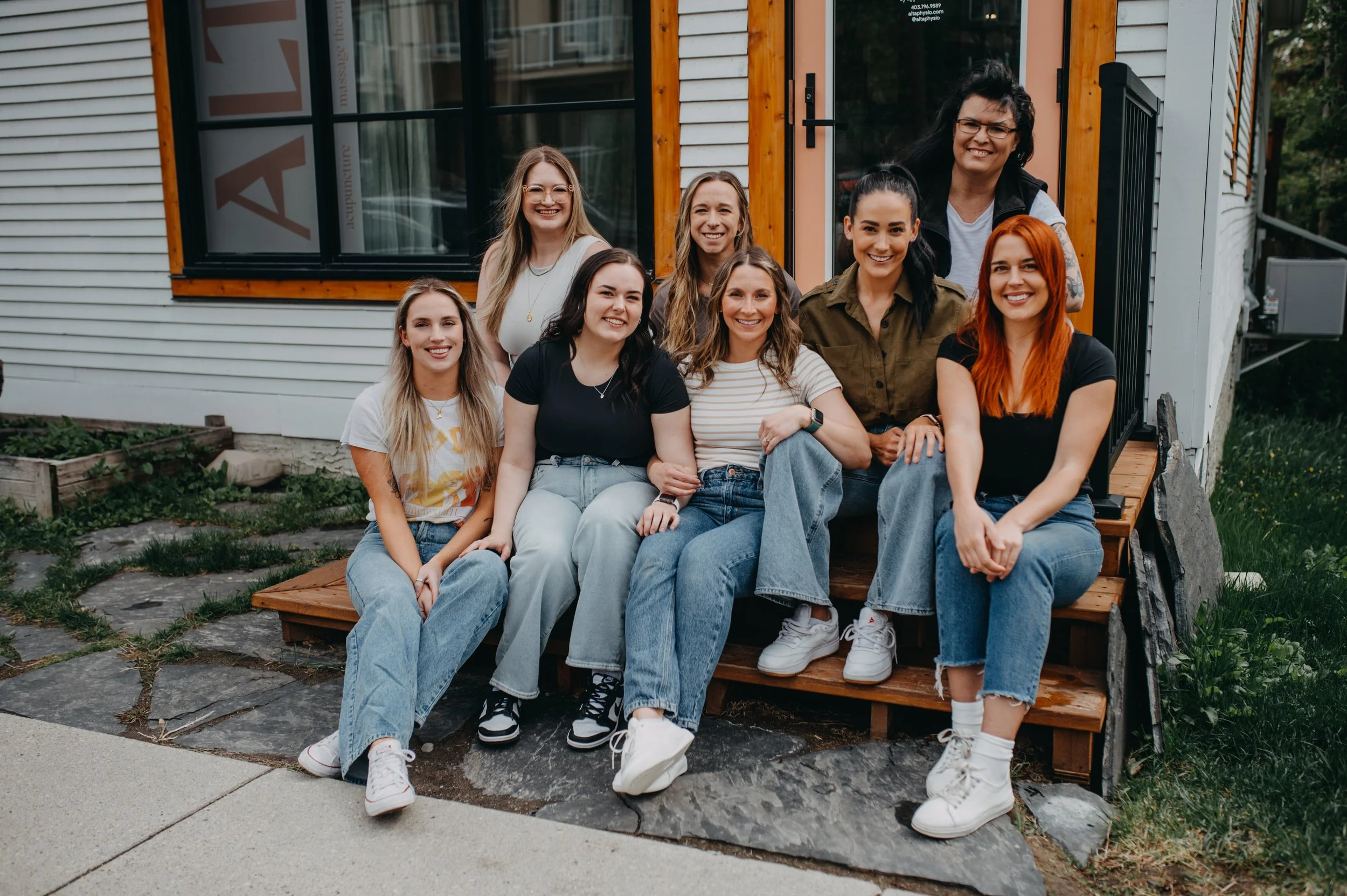 A group of nine diverse women sitting and standing on a wooden porch in front of a house, smiling and posing for a photo.
