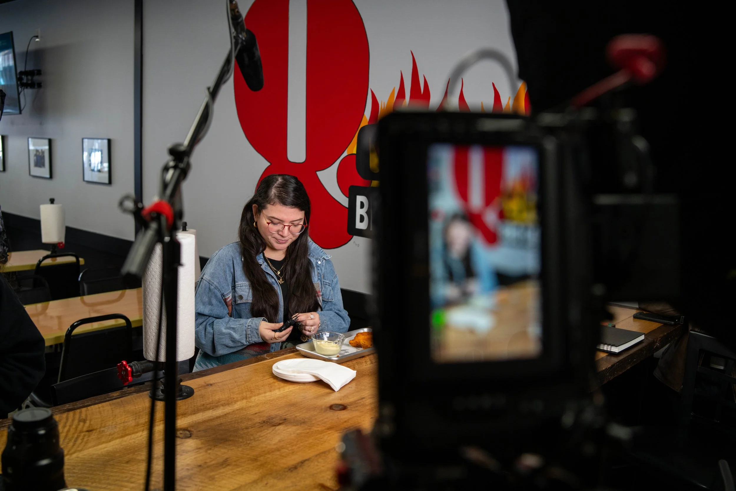 Behind-the-scenes view of a video camera screen capturing talent setting up in a restaurant, with microphone equipment visible during a social media content shoot.