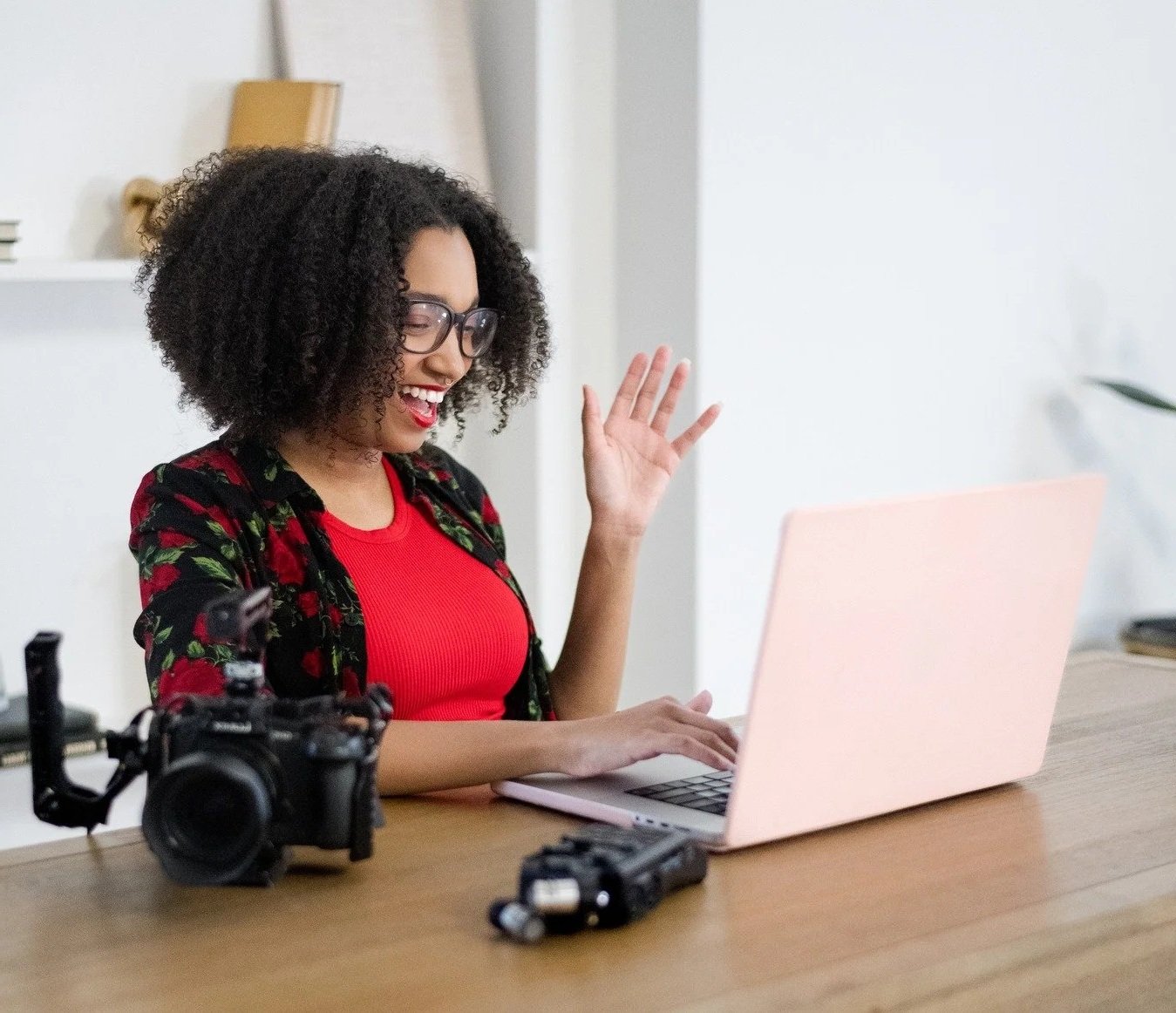 Danielle, founder of FroMotion Media, smiling and waving at her computer during a content strategy session, with photo and video gear on her desk.