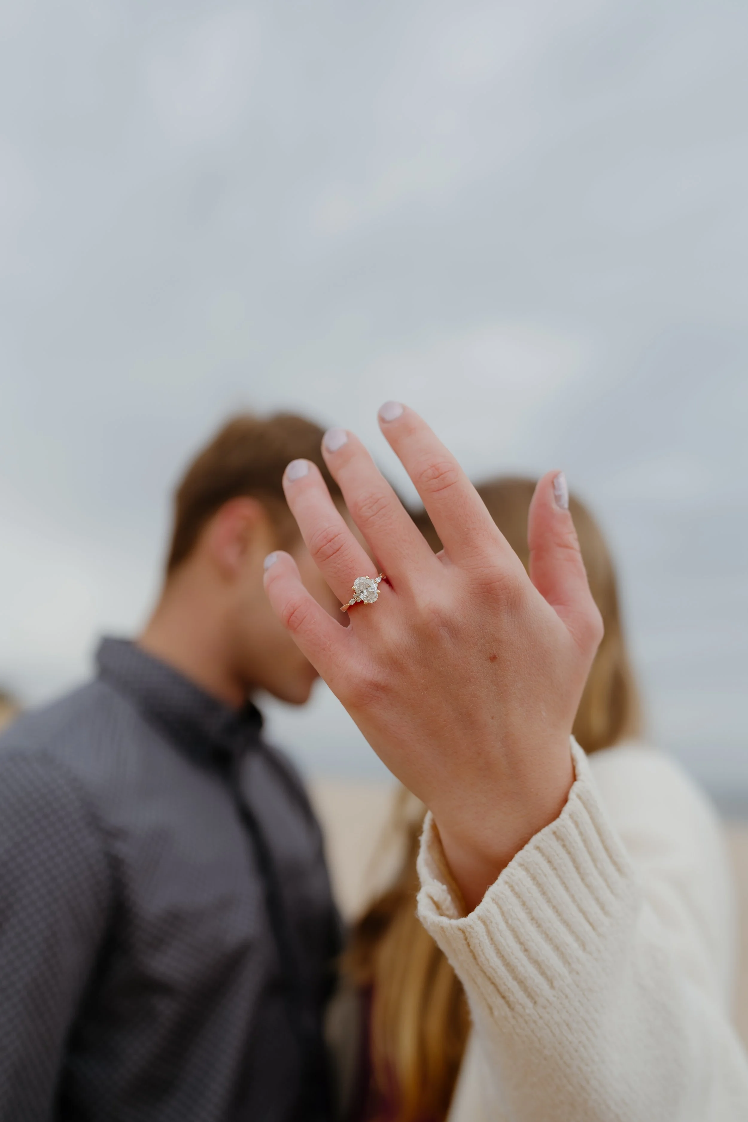 Close-up of a woman's hand showing an engagement ring with a large diamond, with a blurred couple in the background, outdoors on a cloudy day.