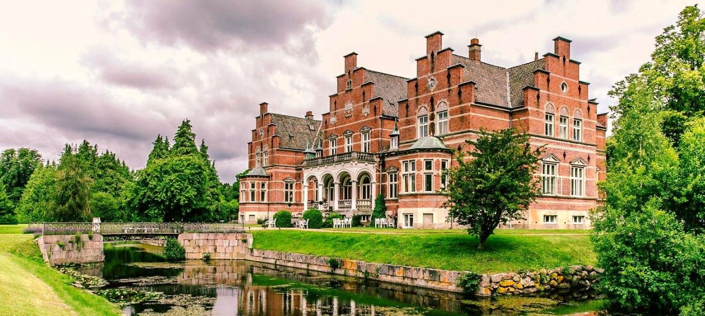 Historisches Gebäude in einem Park mit Fluss, Bäumen und Wolken am Himmel.