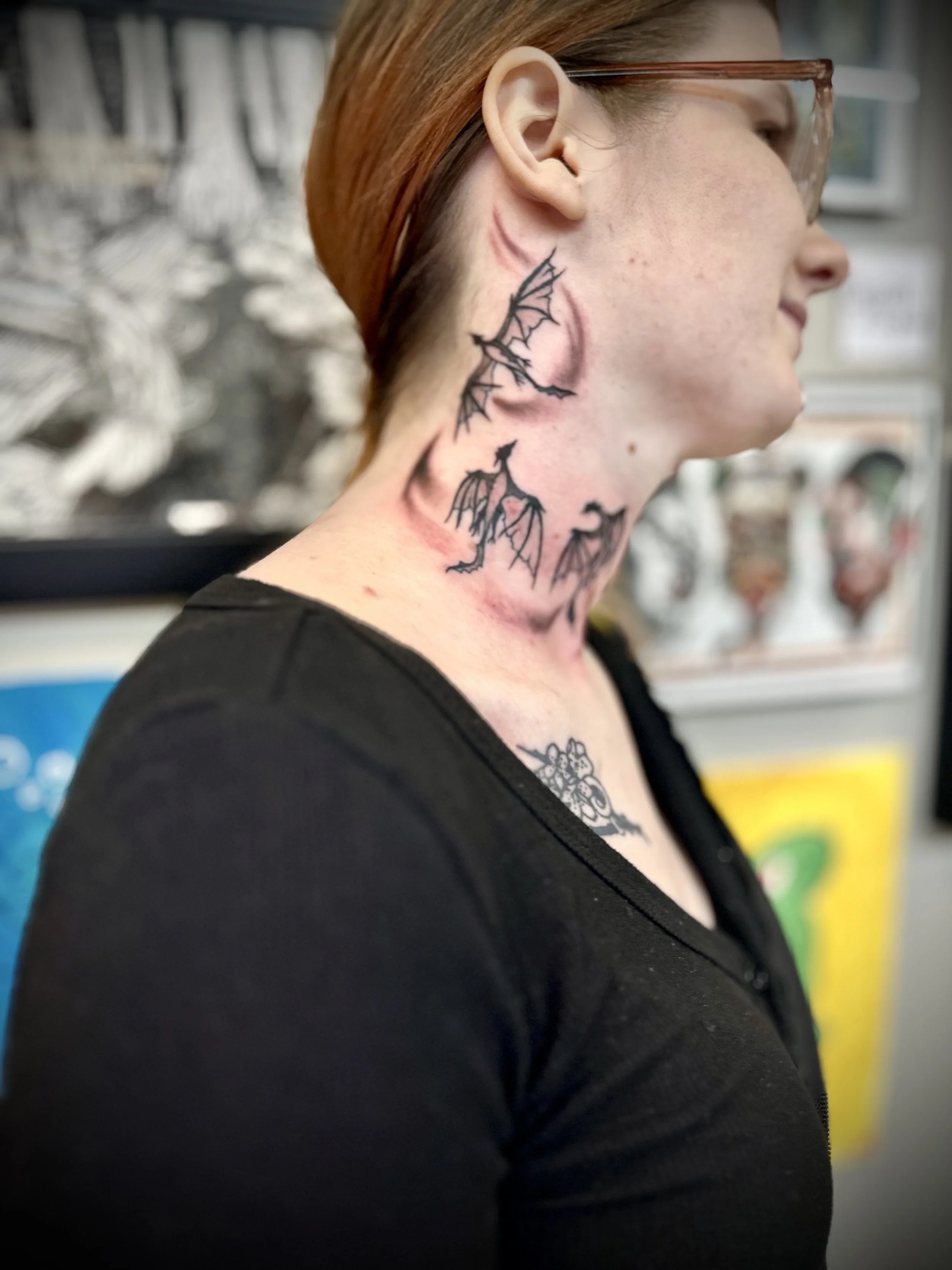 Side profile of a woman with tattoos of bats on her neck, wearing glasses and a black shirt, in an art studio or tattoo shop.