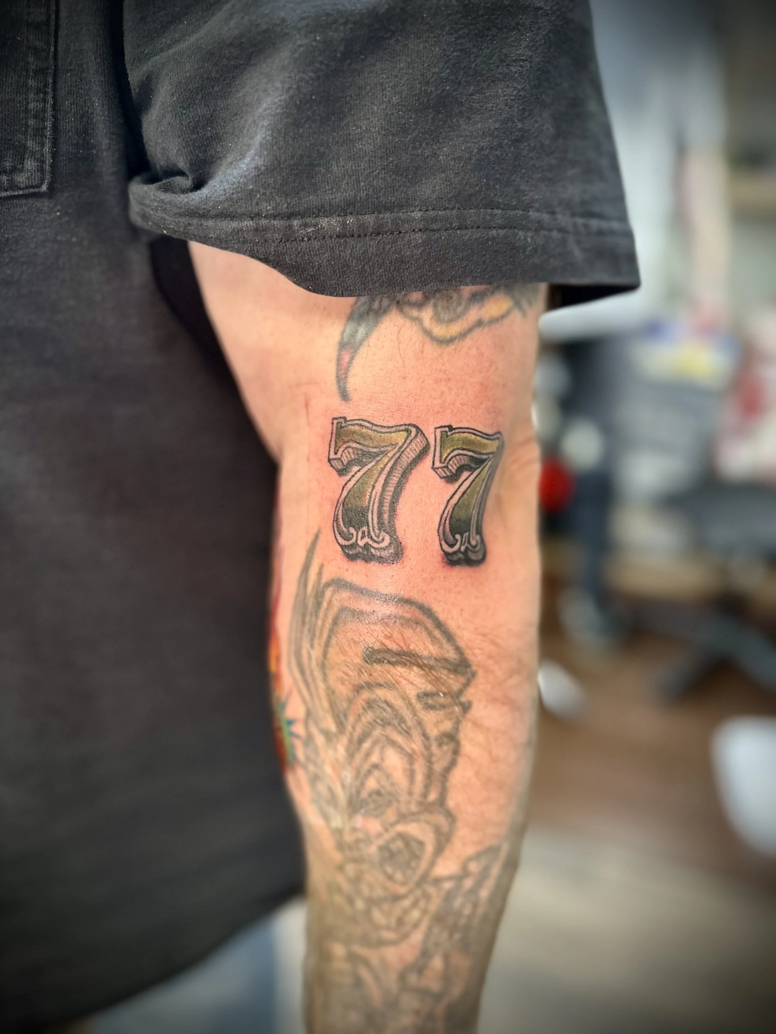Tattooed arm with the number 77 and a dog's face on the forearm, wearing a black t-shirt.