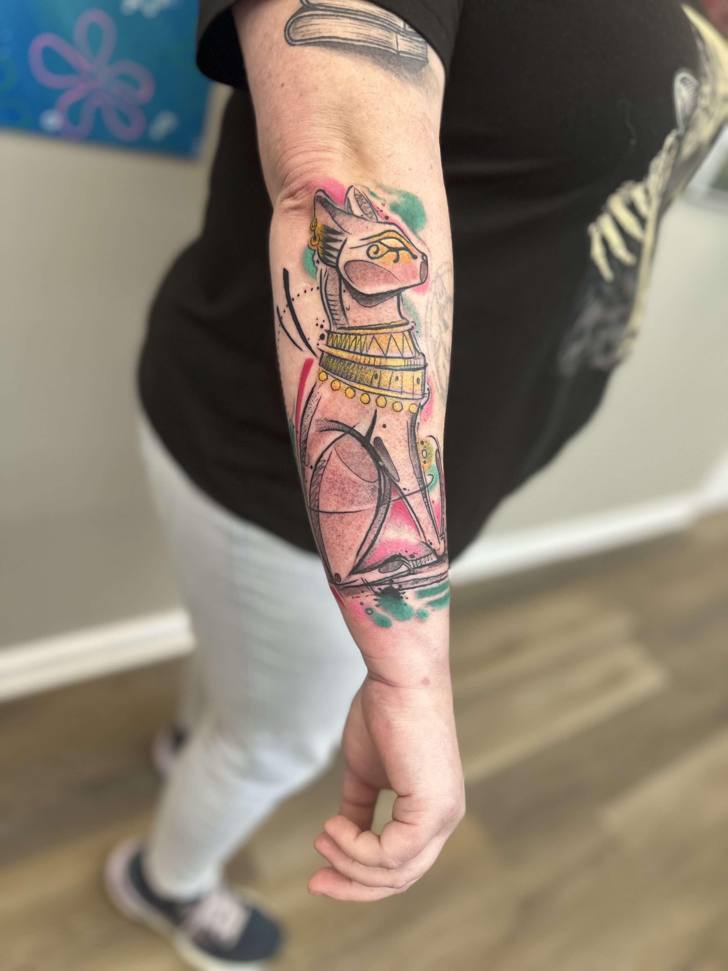 Tattoo of a stylized cat with Egyptian-style jewelry and markings on a person's forearm.