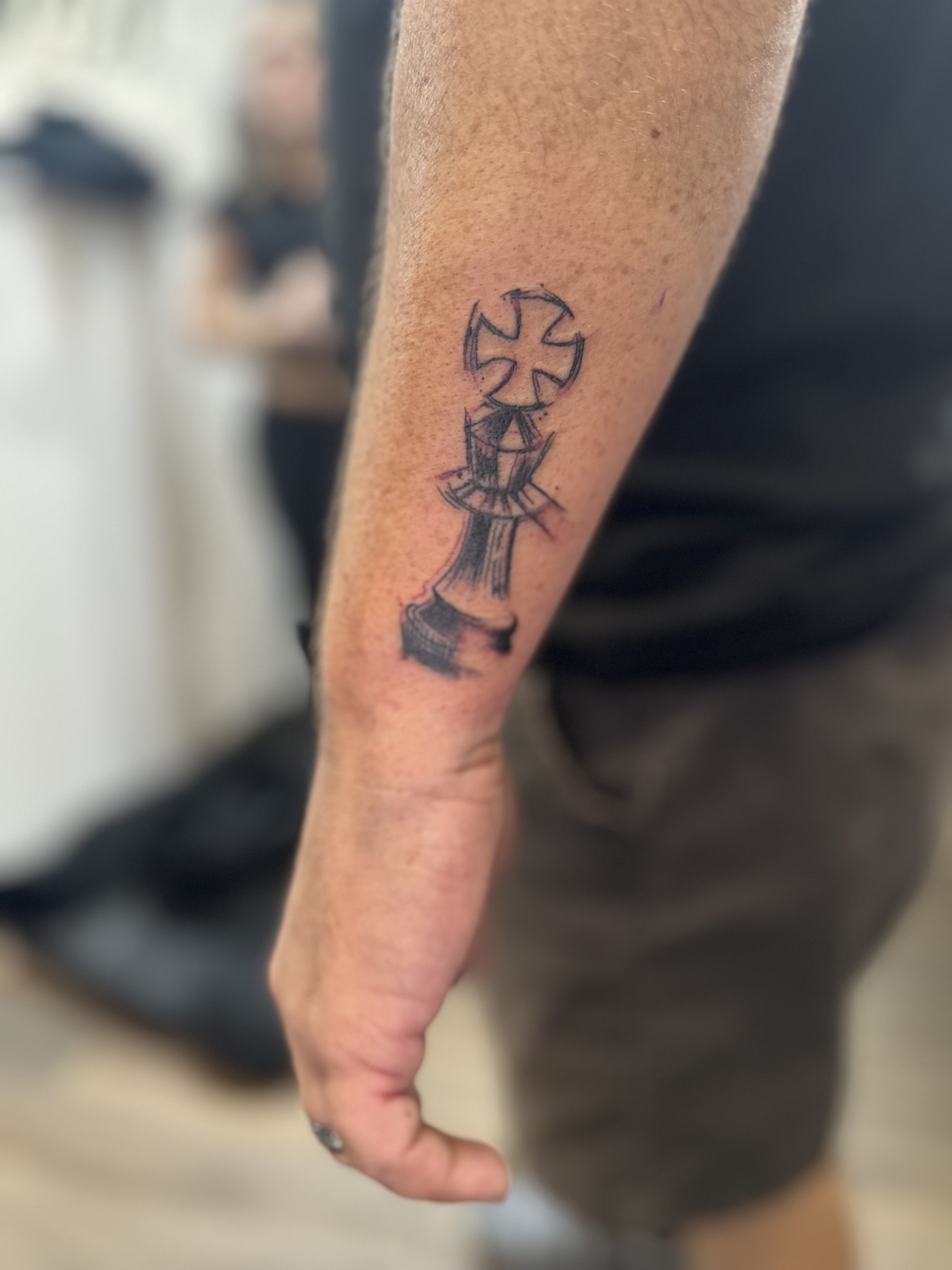 A tattoo of a chess king piece with a stylized cross or kite shape on top, sketched in black ink on a person's forearm.