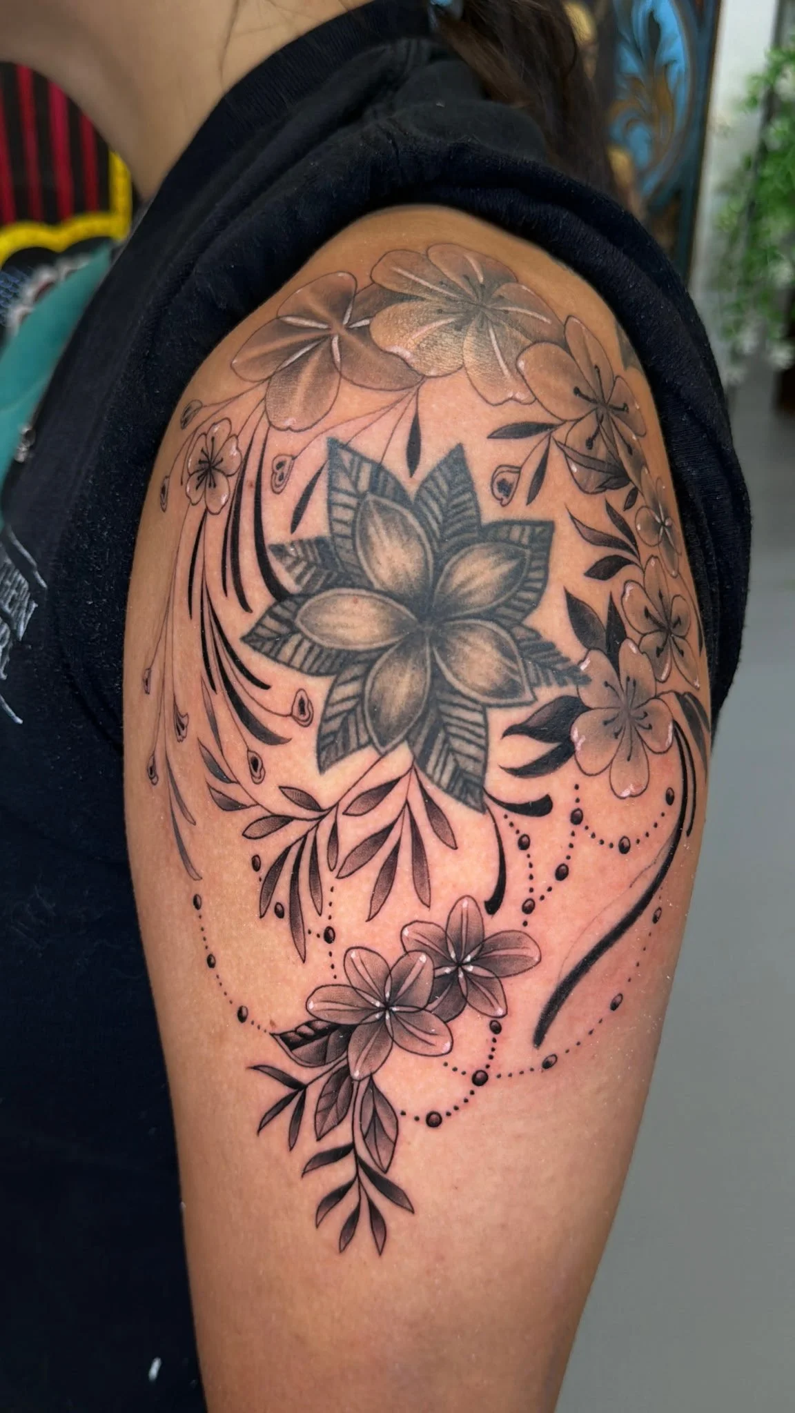Black and gray floral tattoo on upper arm featuring large central flower, smaller surrounding flowers, leaves, and decorative dotted lines.