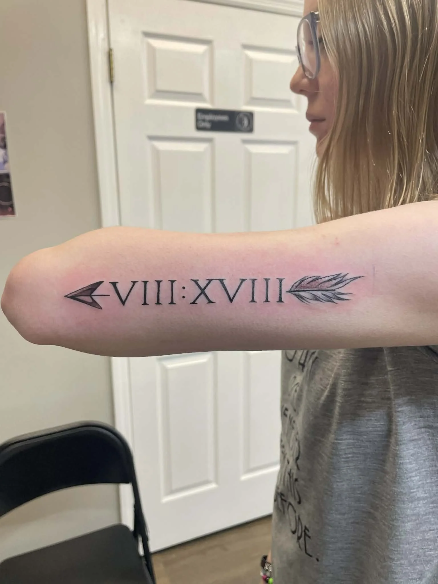 A woman with glasses showing a fresh tattoo of Roman numerals and a feather arrow on her forearm, standing indoors near a white door.