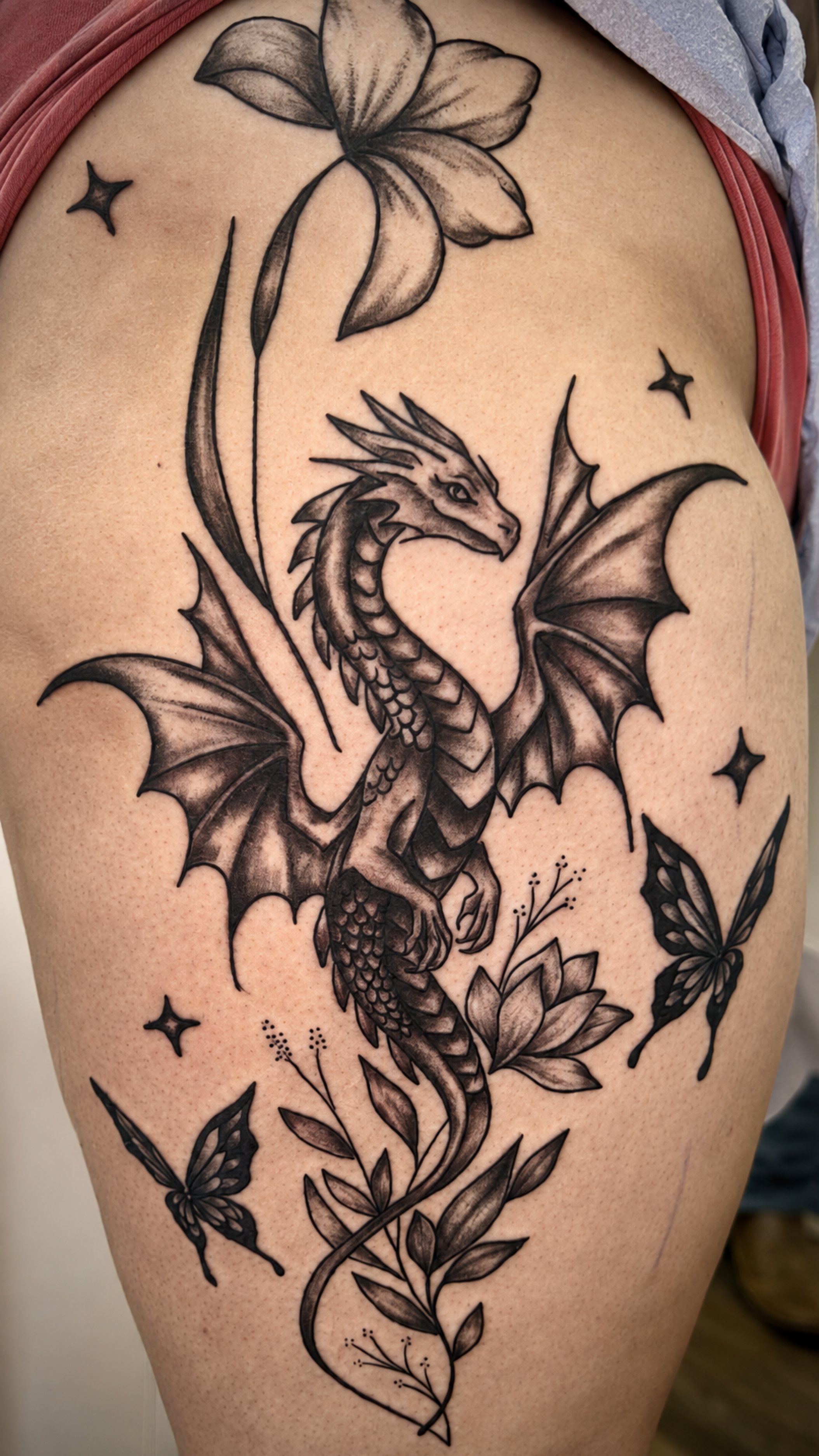 Black tattoo of a dragon with wings, along with flowers, butterflies, stars, and a leaf on skin.
