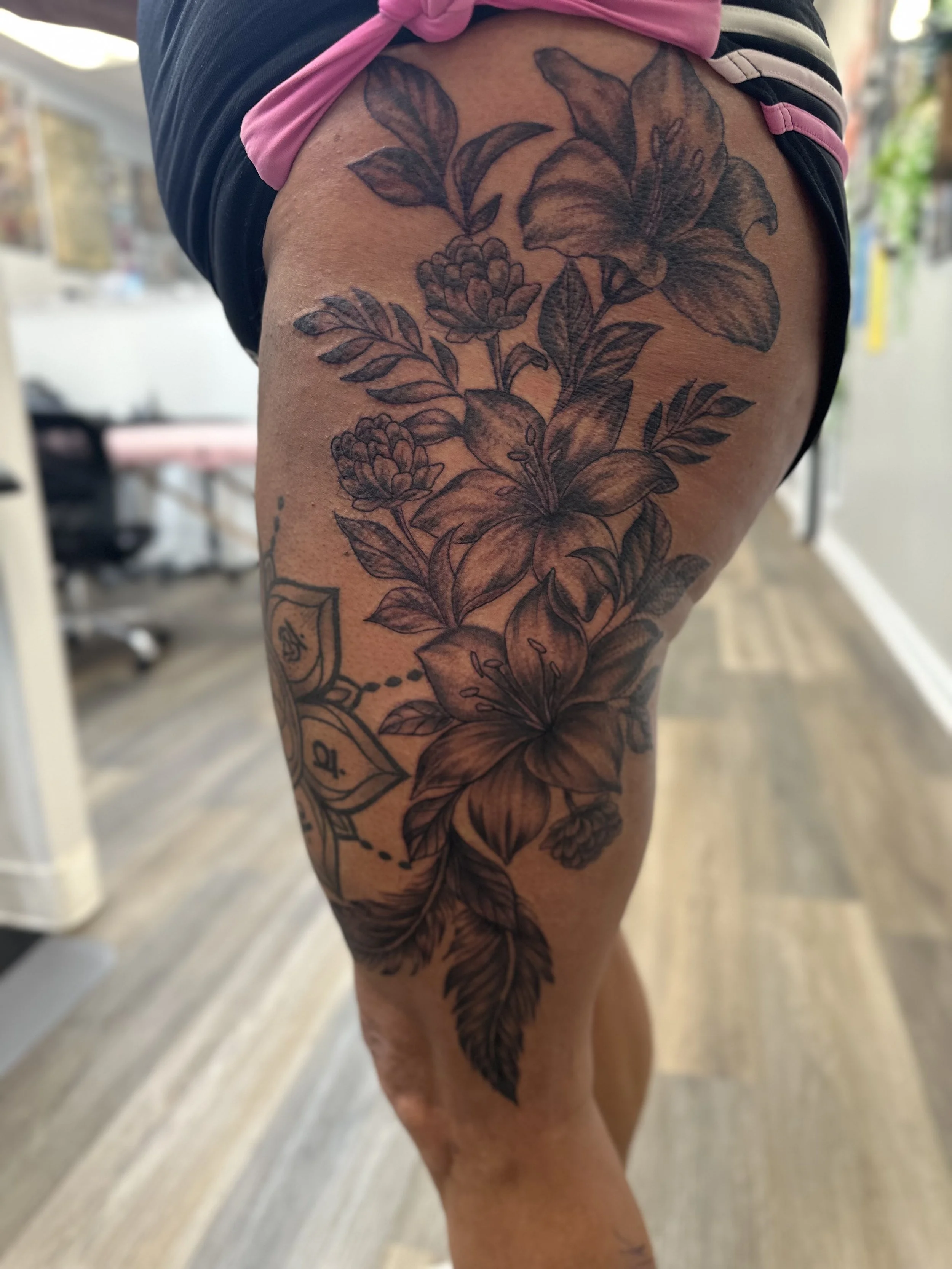 Black ink floral tattoo featuring lilies, roses, and leaves on a person's thigh.