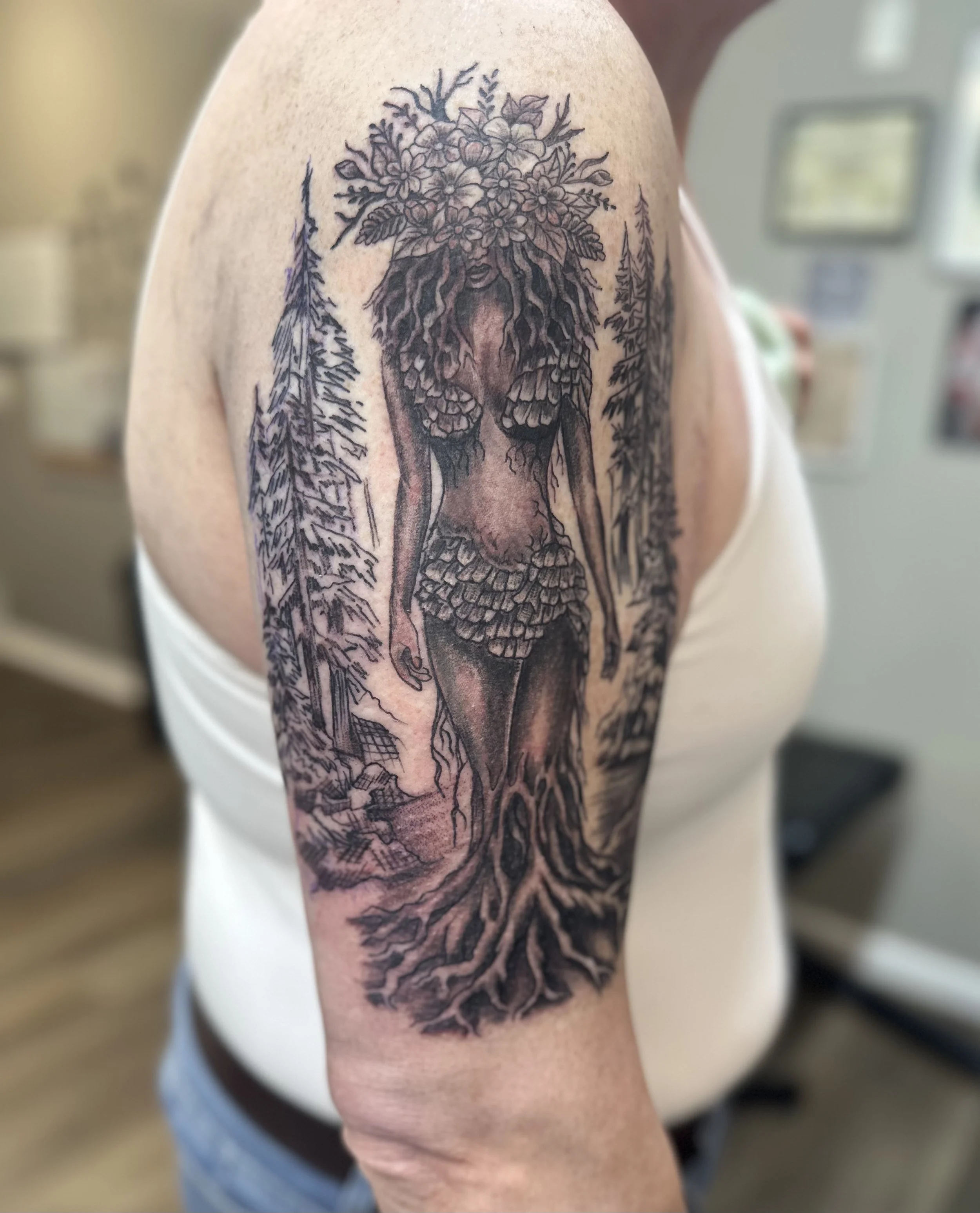 Tattoo of a woman with tree roots as her legs, branches as her arms, and a floral crown on her head, surrounded by pine trees.
