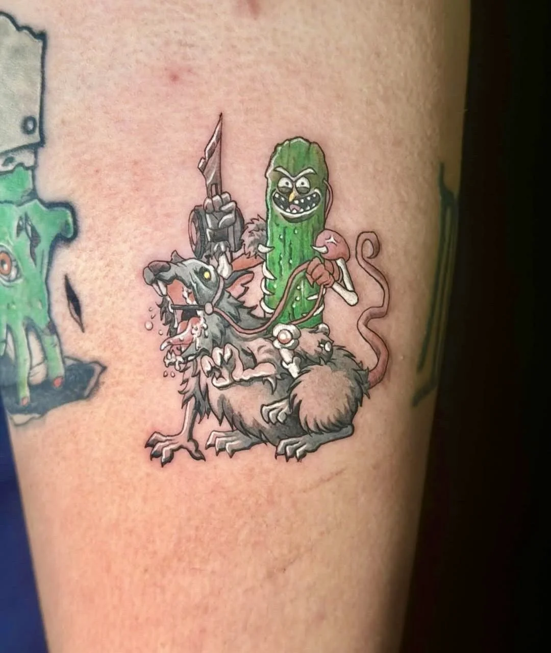 A cartoon-style tattoo of a rat with a pickle head riding on a furry animal, holding a sword and a pink leash, with a pink tongue and sharp teeth, on a person's skin.