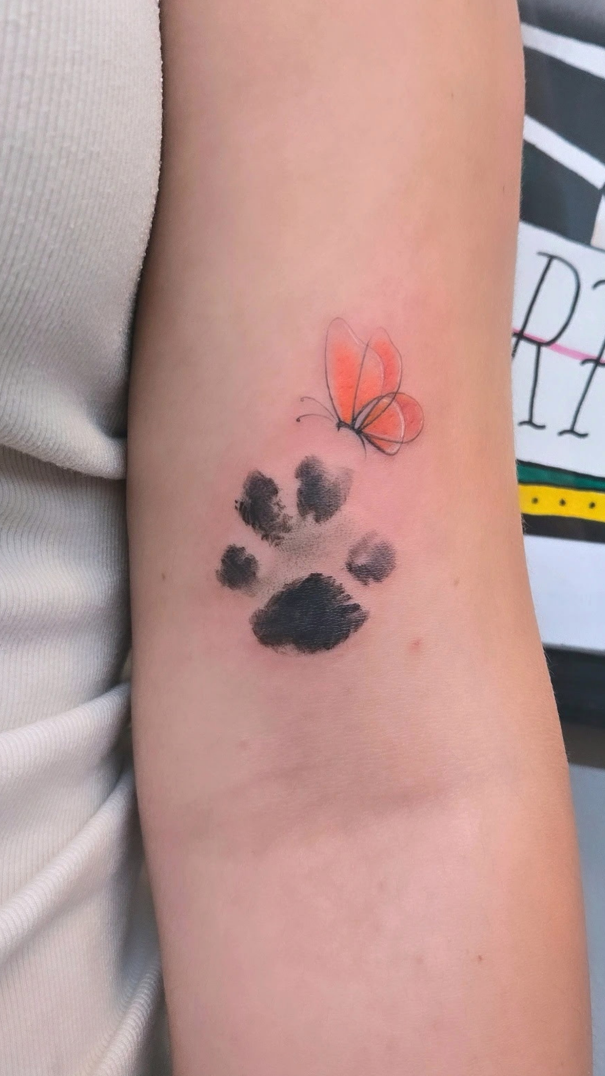 Tattoo of a butterfly with orange wings above a black paw print on a person's arm.
