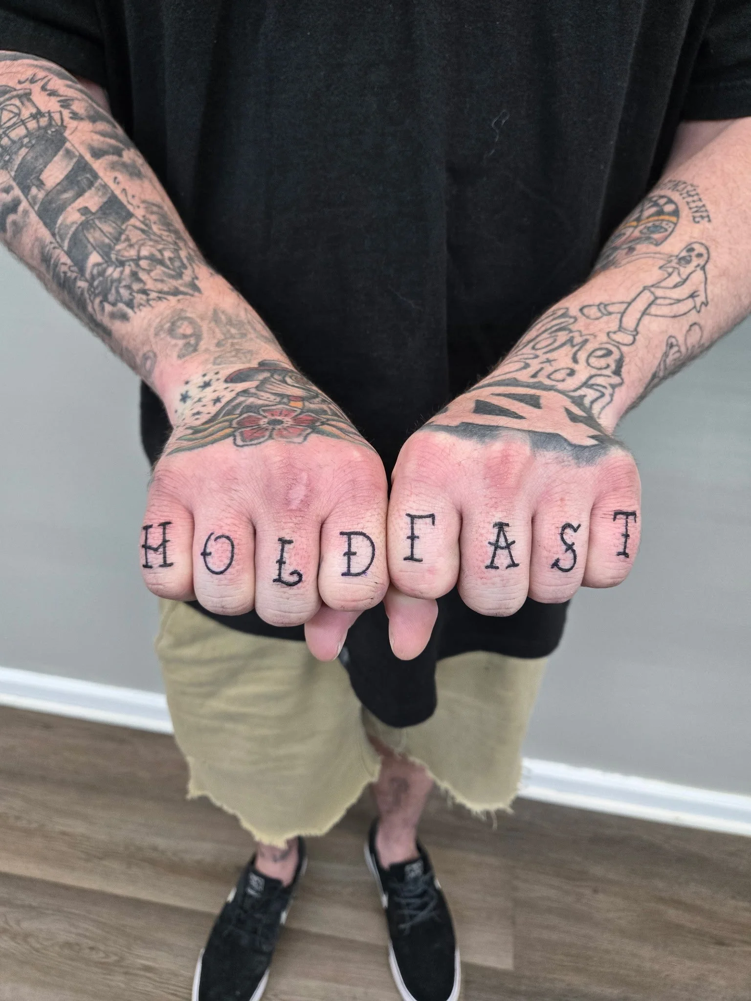 Tattooed man with clenched fists displaying the word 'HOLD FAST' tattooed on knuckles, standing indoors on a wooden floor, wearing a black shirt, beige shorts, and black sneakers.