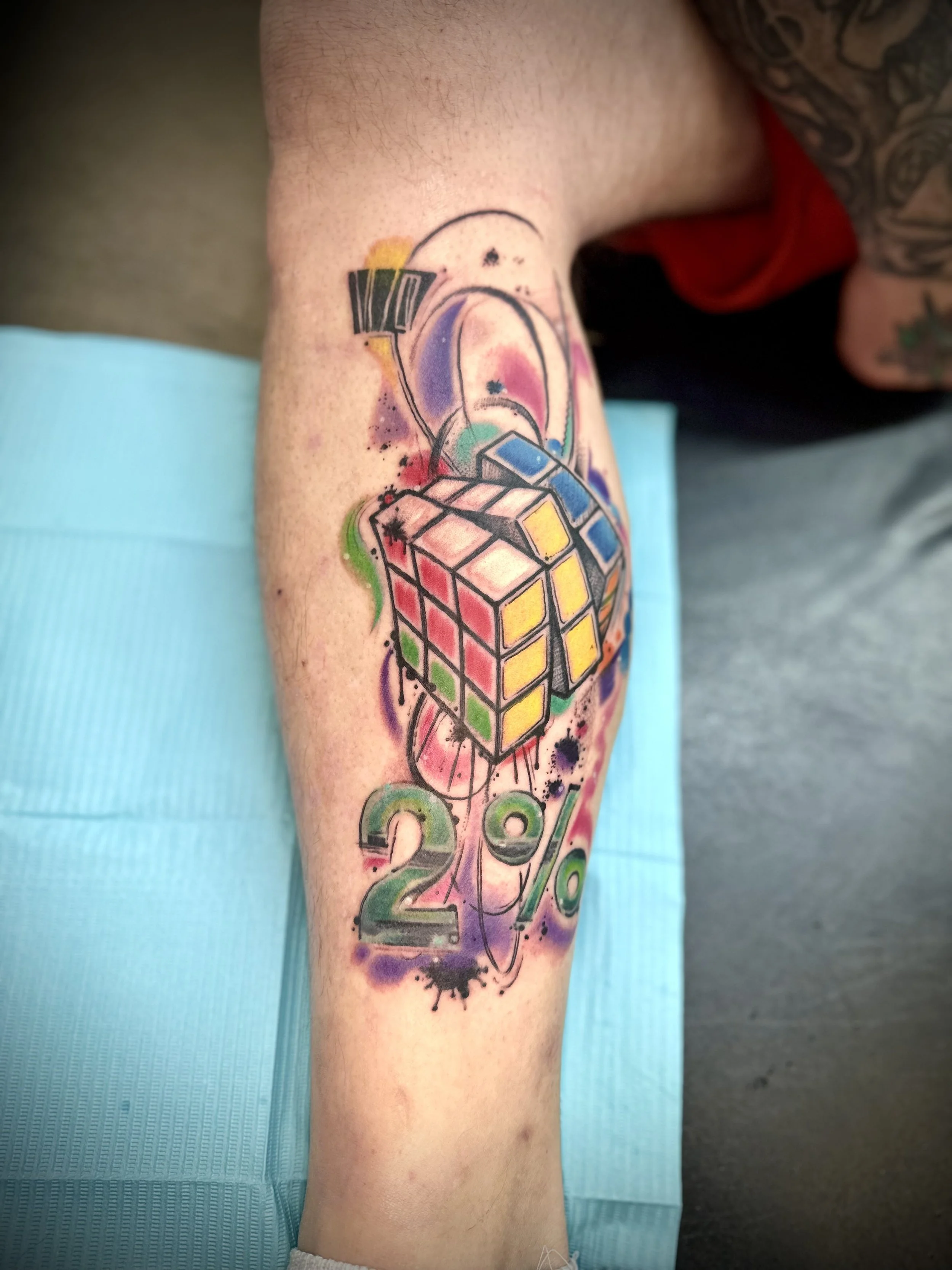 Colorful tattoo of a Rubik's cube, a 2% symbol, and abstract splashes and lines on a person's arm.