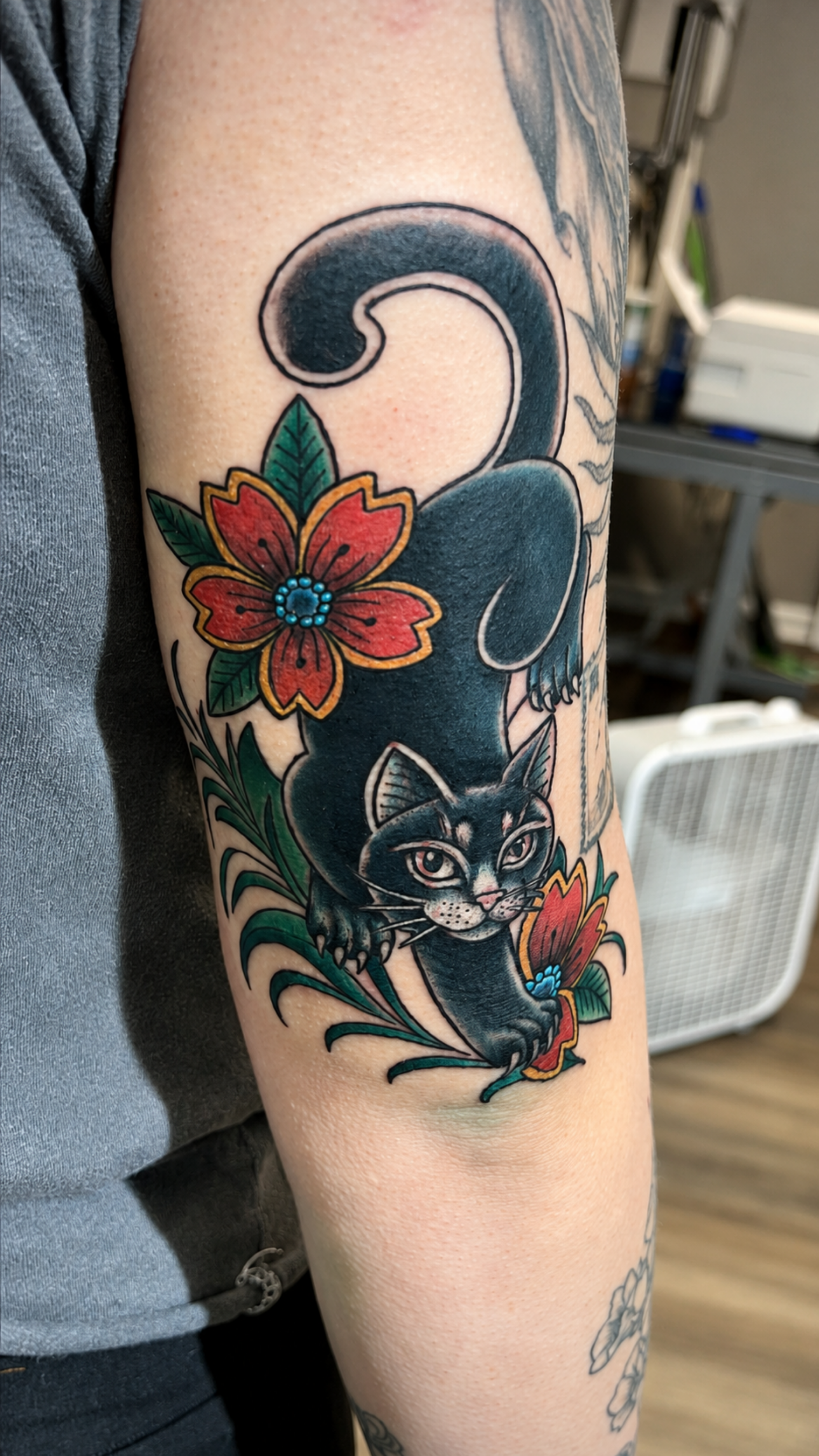 Colorful tattoo on person's upper arm featuring a black cat with yellow eyes, surrounded by red flowers with green leaves, and a large black spiral shape at the top.