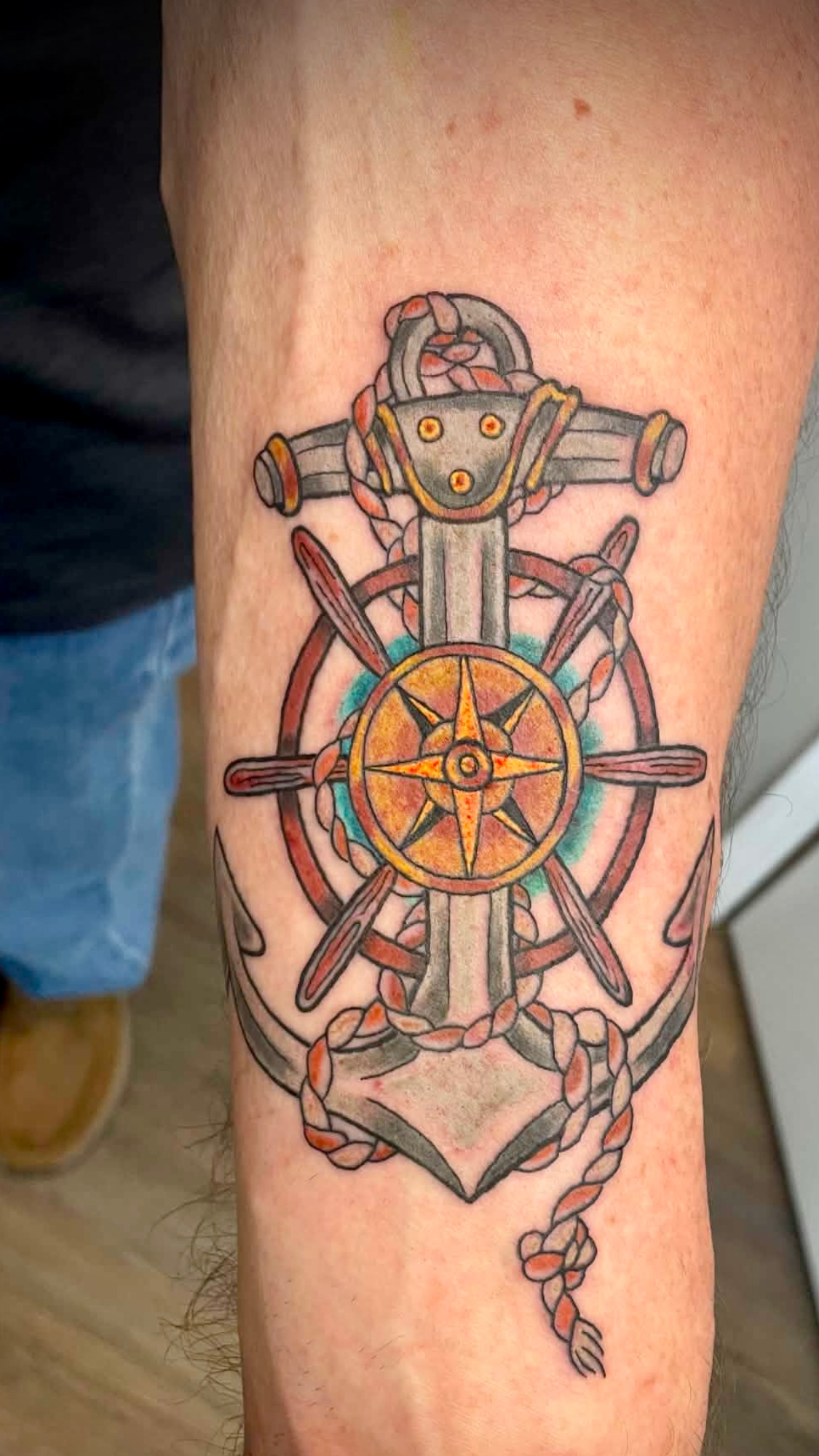 Tattoo of a ship's steering wheel with a rope wrapped around it on a person's arm.