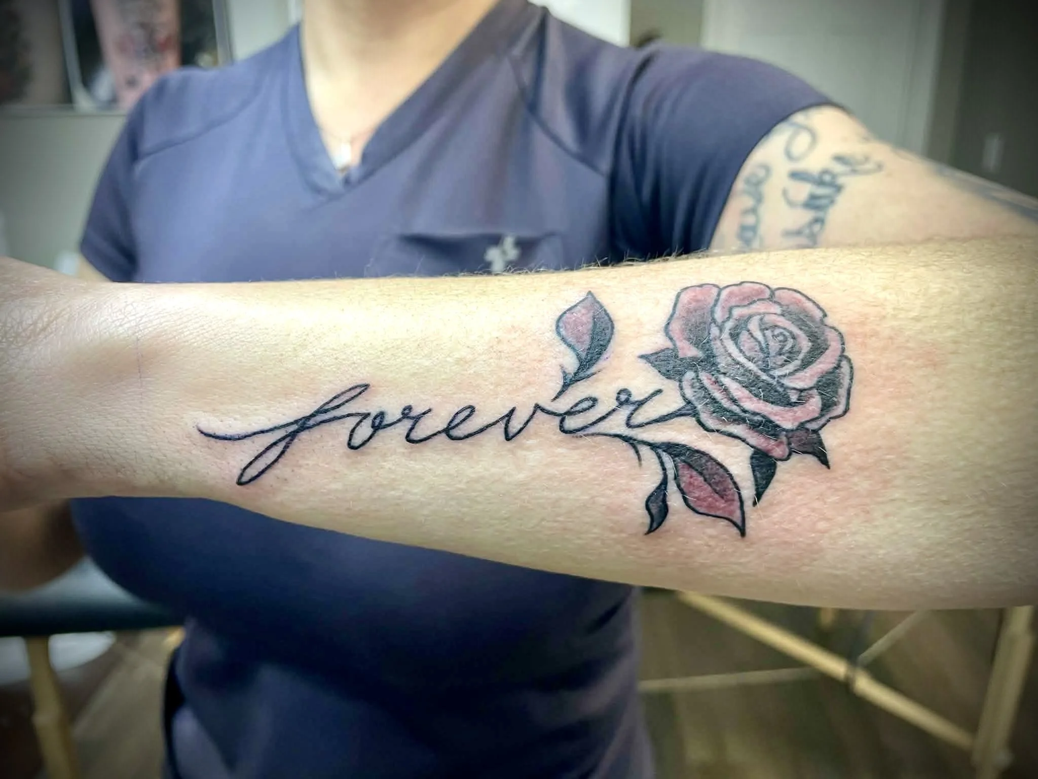 A person's forearm with a tattoo of the word 'forever' in script alongside a pink and black rose with green leaves.