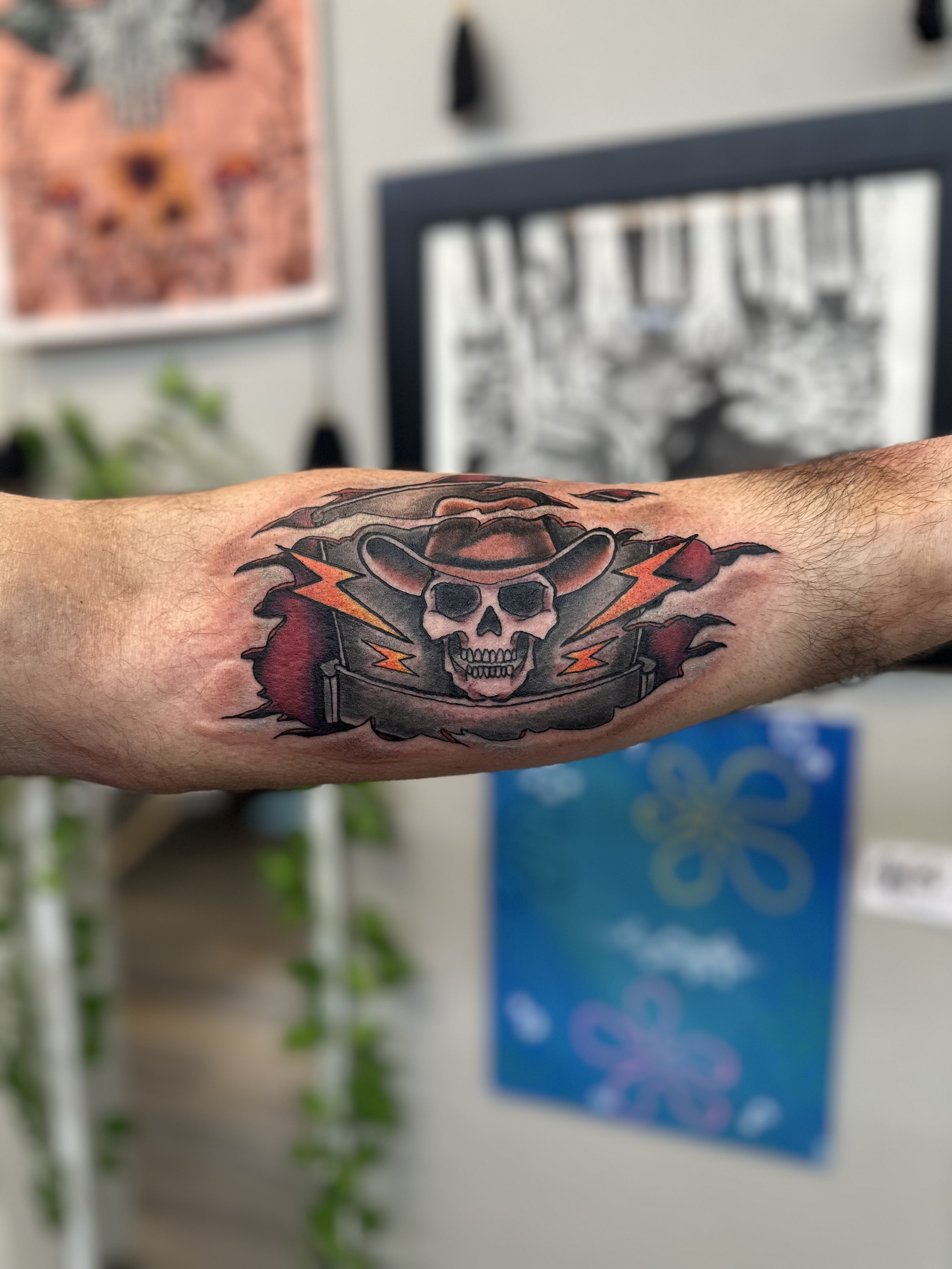 Tattoo of a skull wearing a cowboy hat with lightning bolts behind it on a person's forearm.