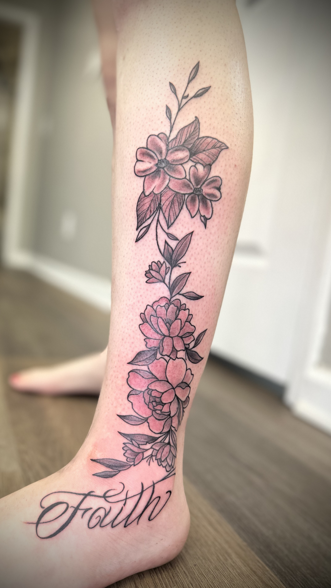 A person's lower leg with a floral tattoo extending from the ankle to mid-calf, featuring pink flowers, green leaves, and the word 'Faith' in script.