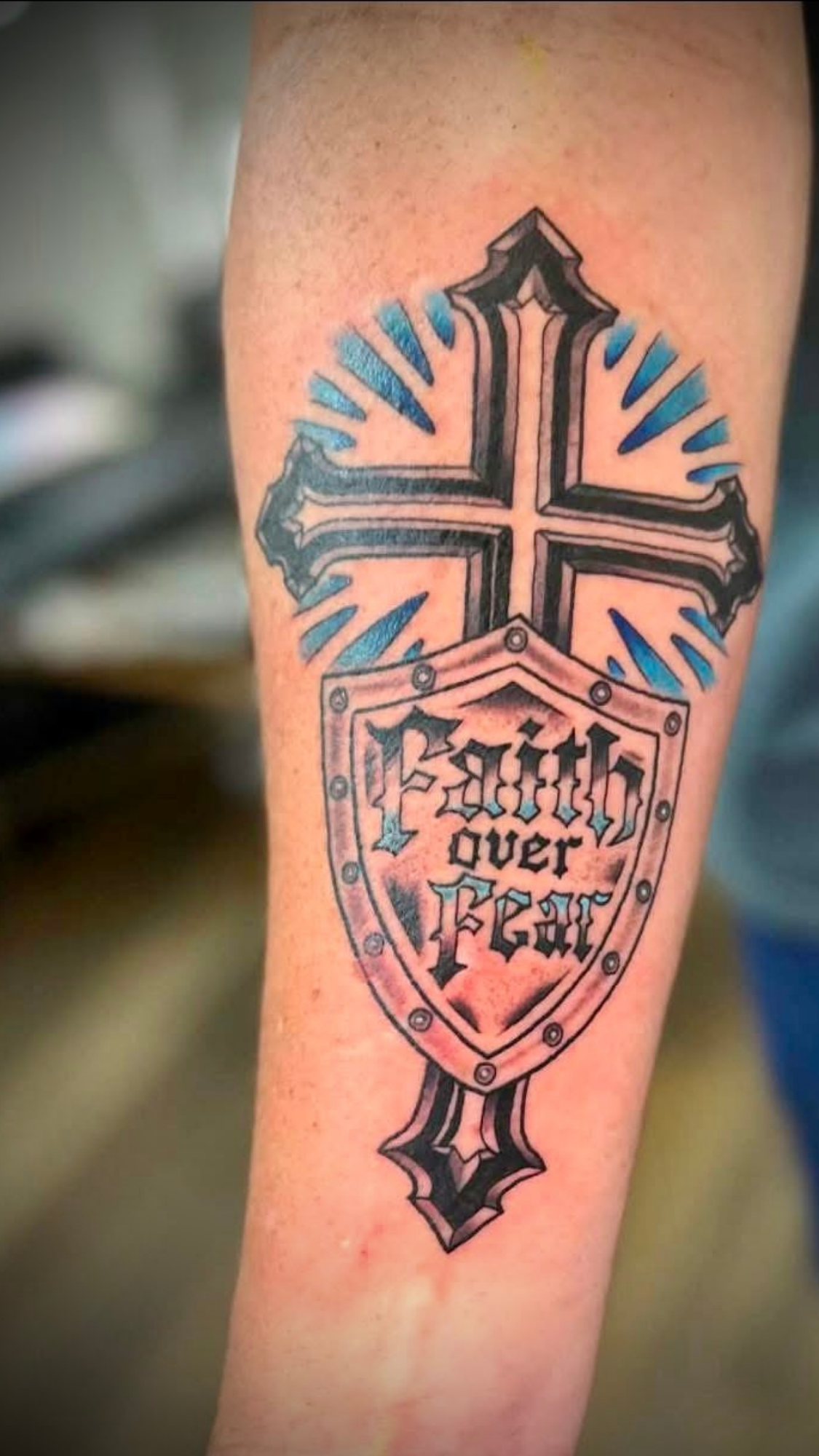 Tattoo of a sword with a cross and a shield with the words "Blood, Over, Fear" on the arm.