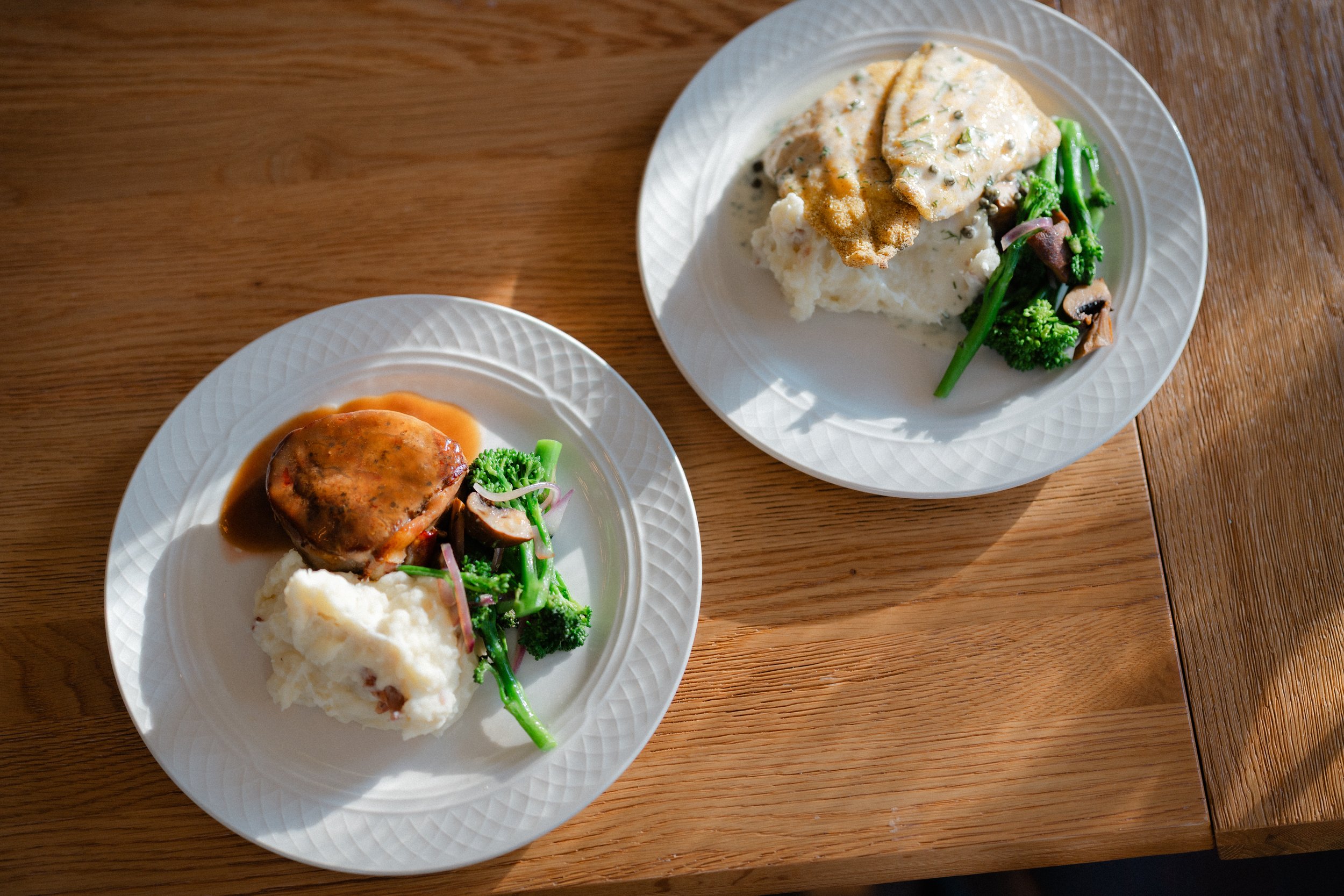 Two white plates of food on a wooden table, each containing mashed potatoes, green vegetables, and a piece of meat with sauce.