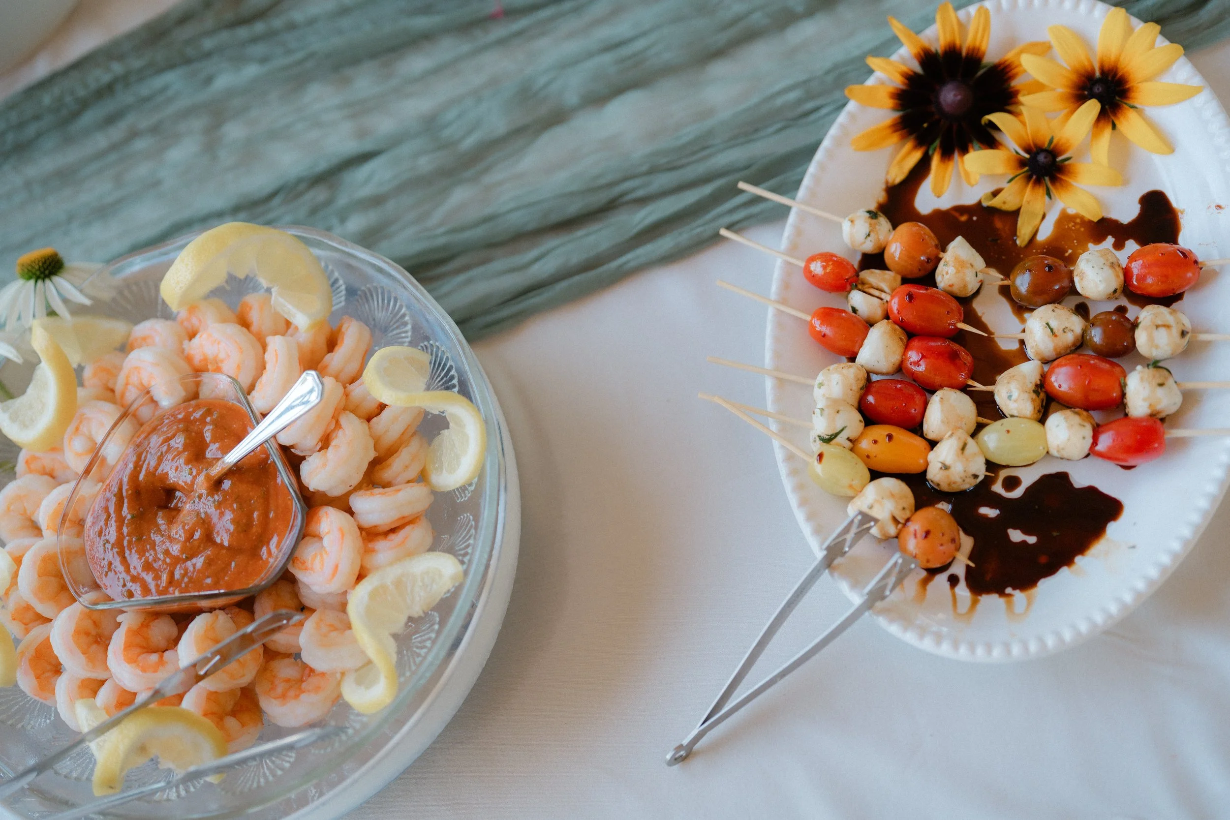 Tray with cooked shrimp and lemon slices with a dipping sauce, and another plate with skewers of various cheese, cherry tomatoes, and garnished with yellow flowers, on a blue-green fabric background.