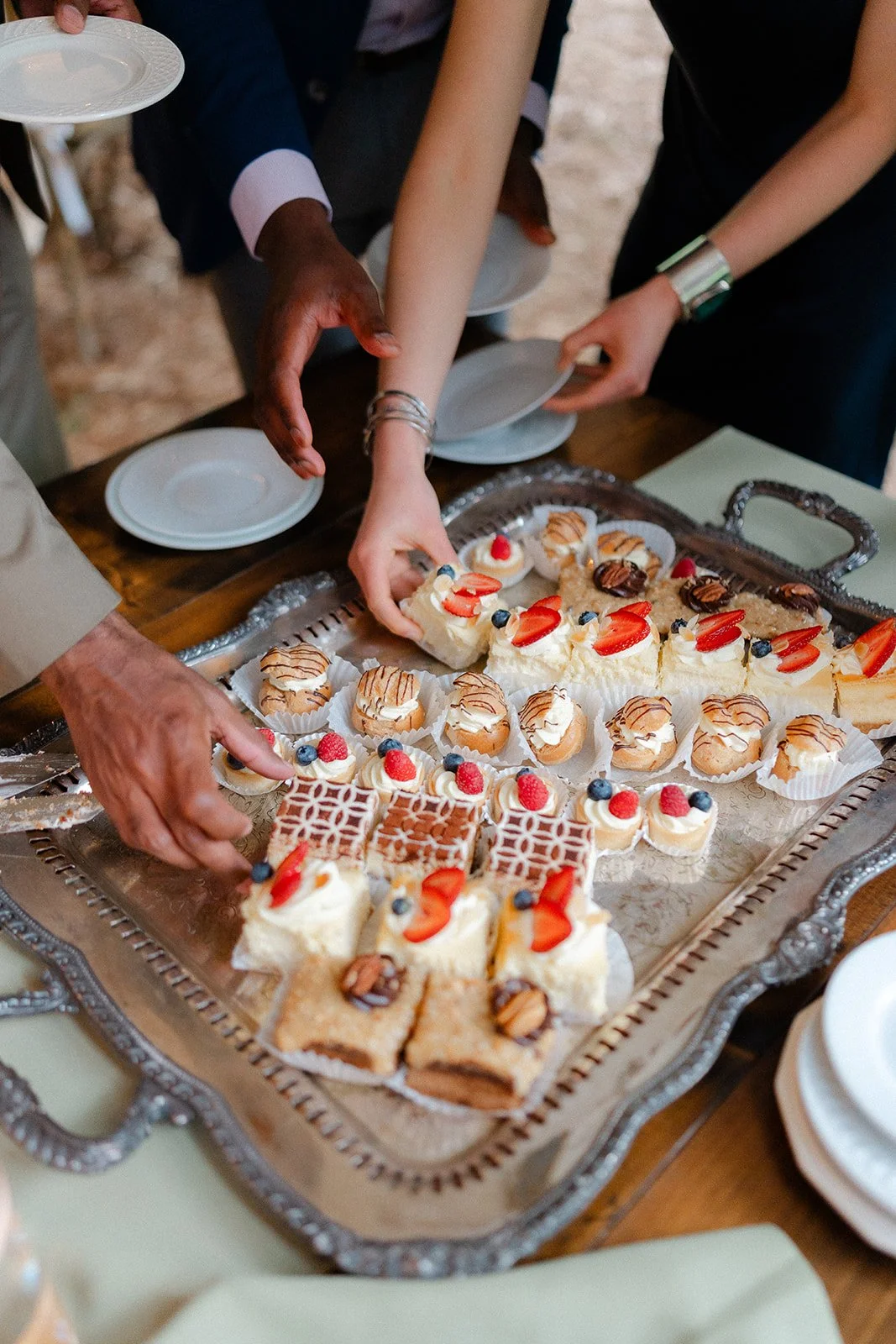 People reaching for an assortment of desserts including small cakes topped with fruit, chocolate, and whipped cream, arranged on a silver tray.