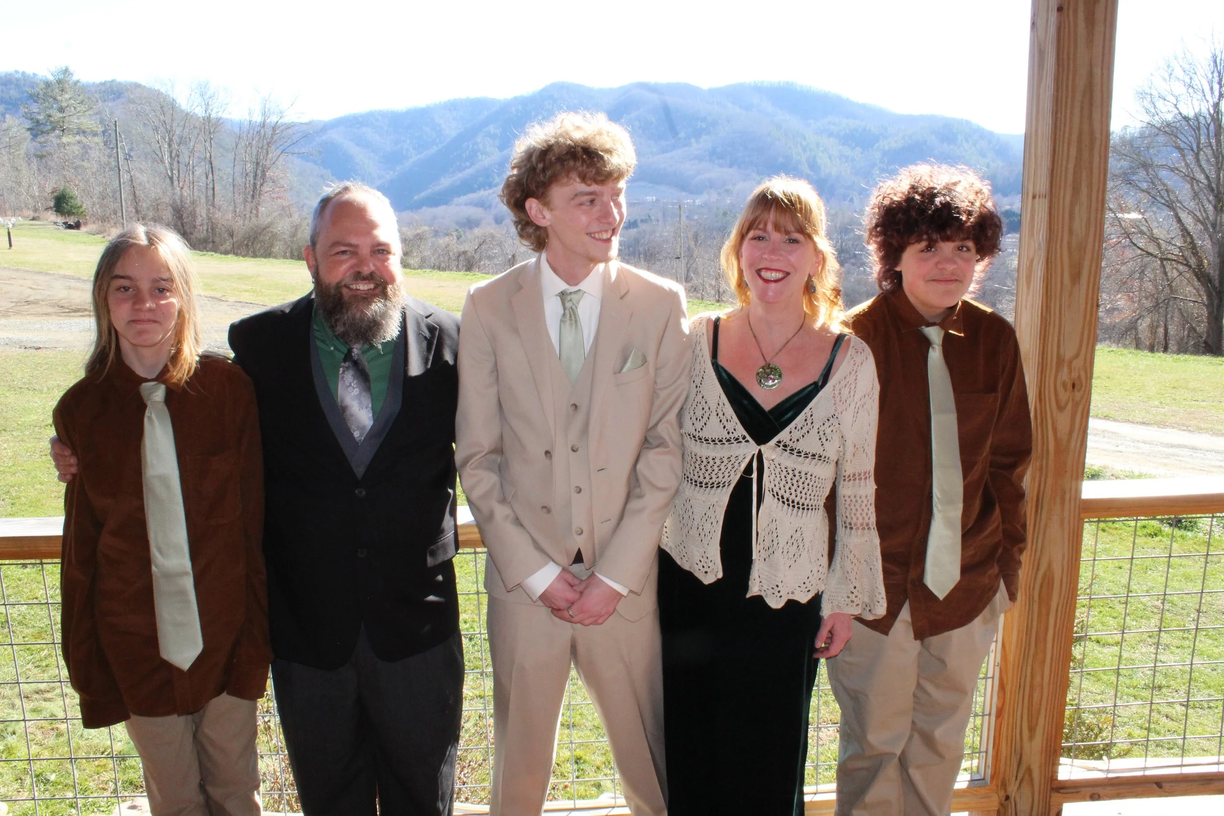 Group of five people standing outdoors on a porch with scenic mountains in the background, smiling and dressed in semi-formal attire.