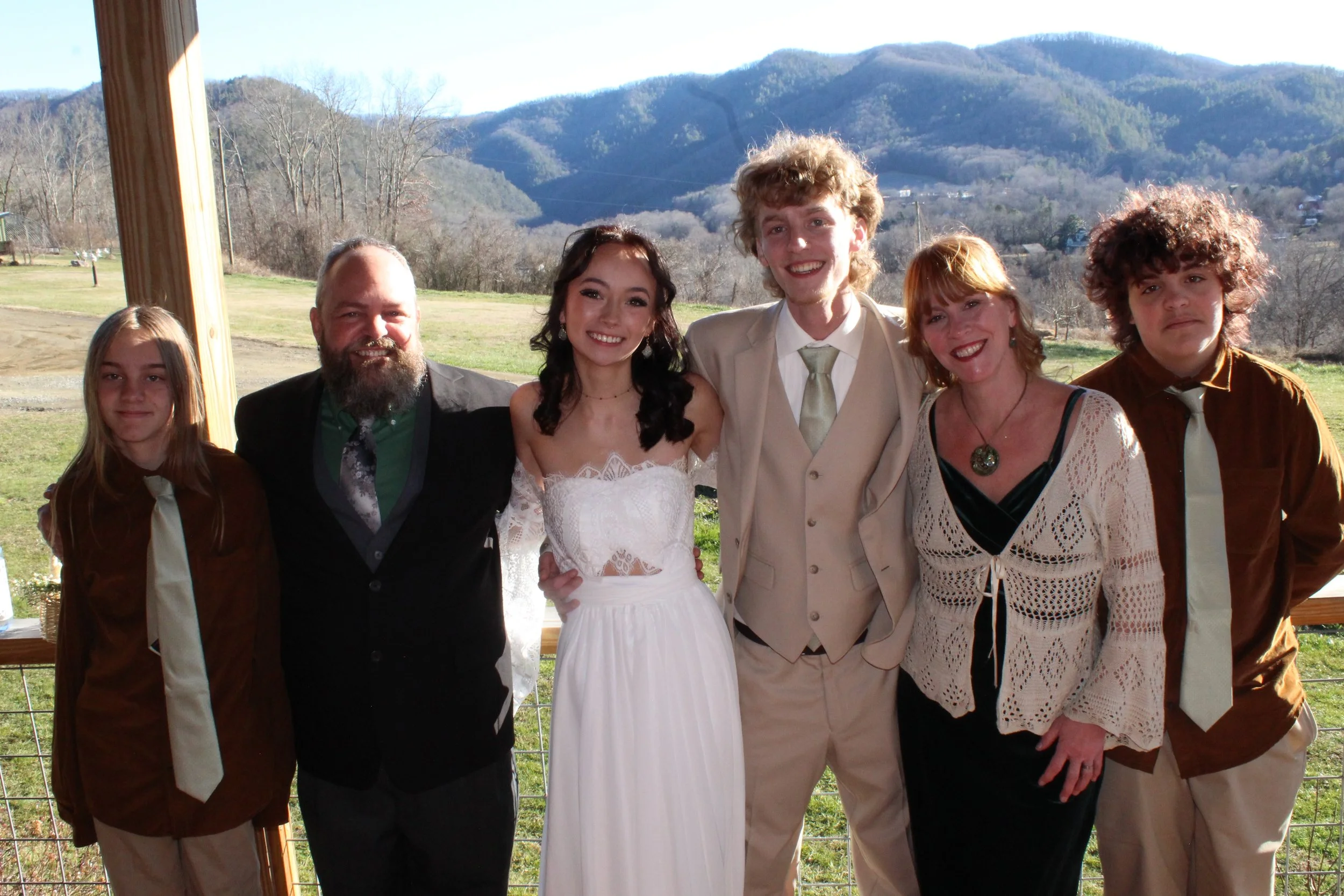 Group of seven people, including one bride and one groom, standing outside with mountains in the background, smiling.