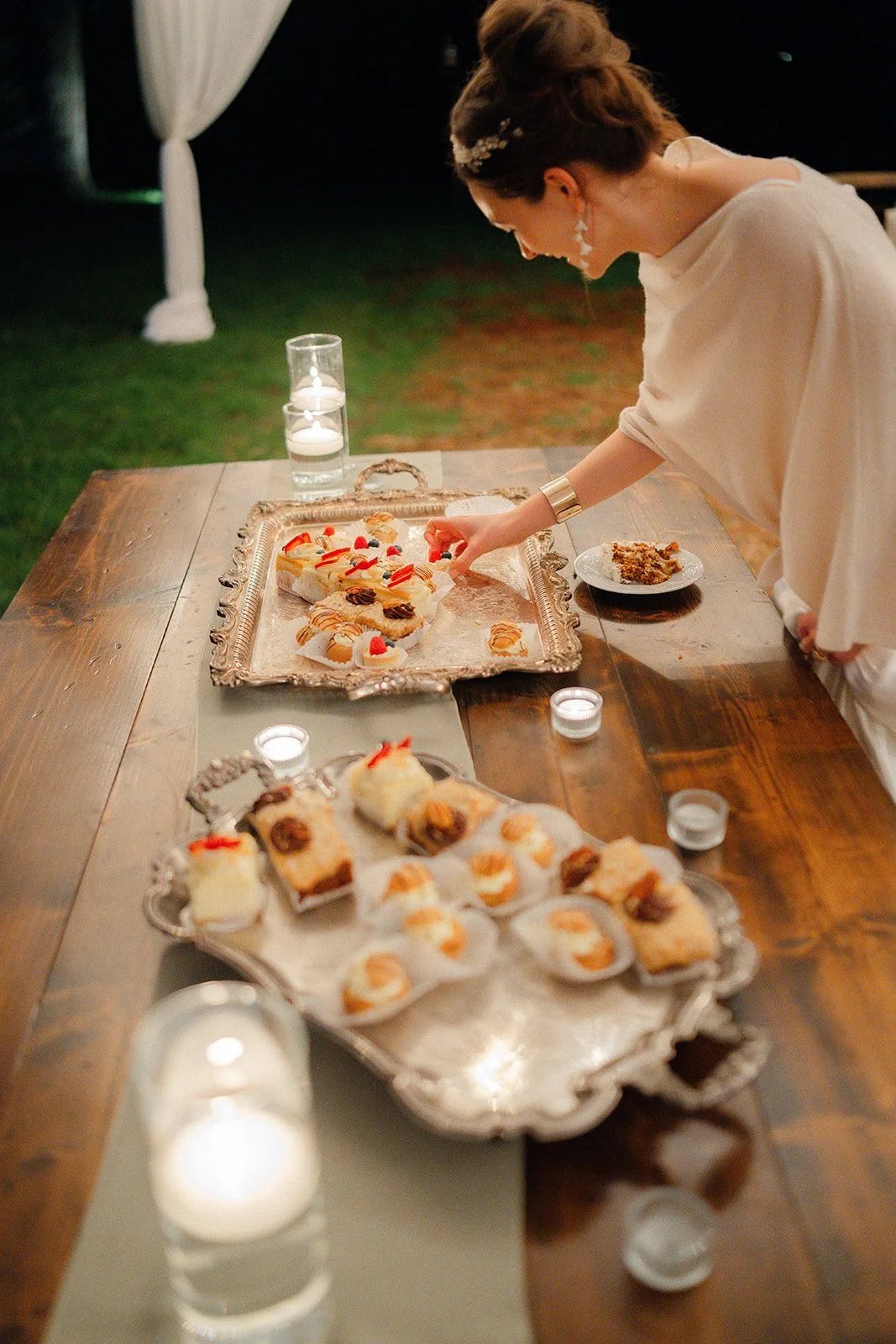 A woman in white is arranging dessert on a silver tray at an outdoor evening event, with candles on the table and a plate of dessert nearby.