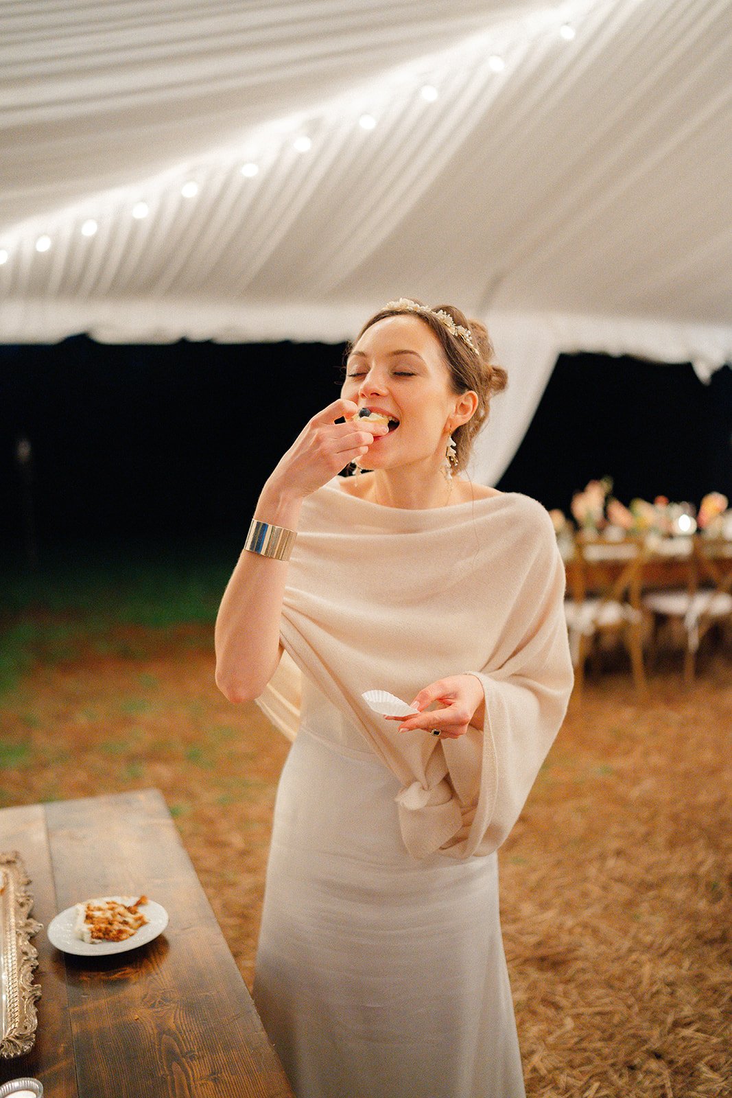 A woman in a cream-colored dress and shawl, wearing a floral headband and earrings, eats a slice of cake at a reception under a white tent with string lights.