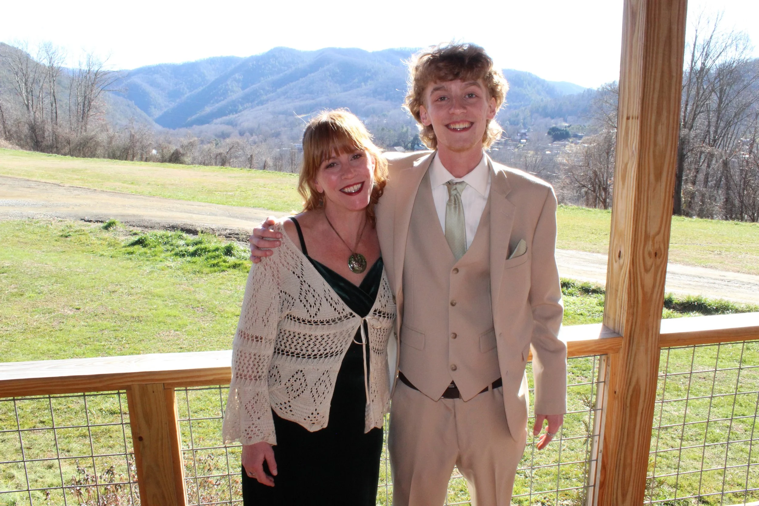 A woman and a man in formal attire standing on a porch with mountains and trees in the background.
