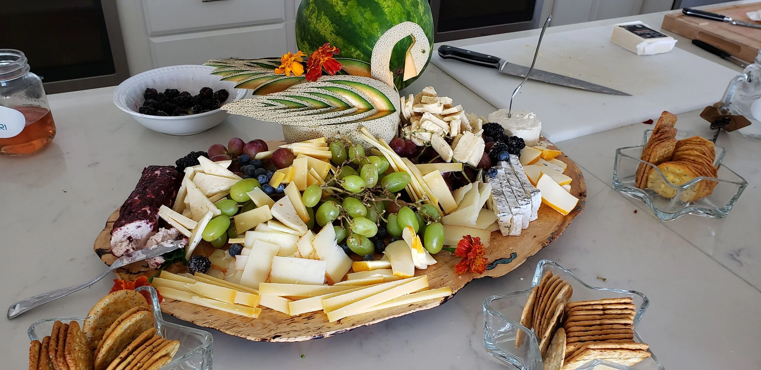 A cheese and fruit platter on a wooden board with grapes, berries, and cheese surrounded by crackers and cookies on a table.