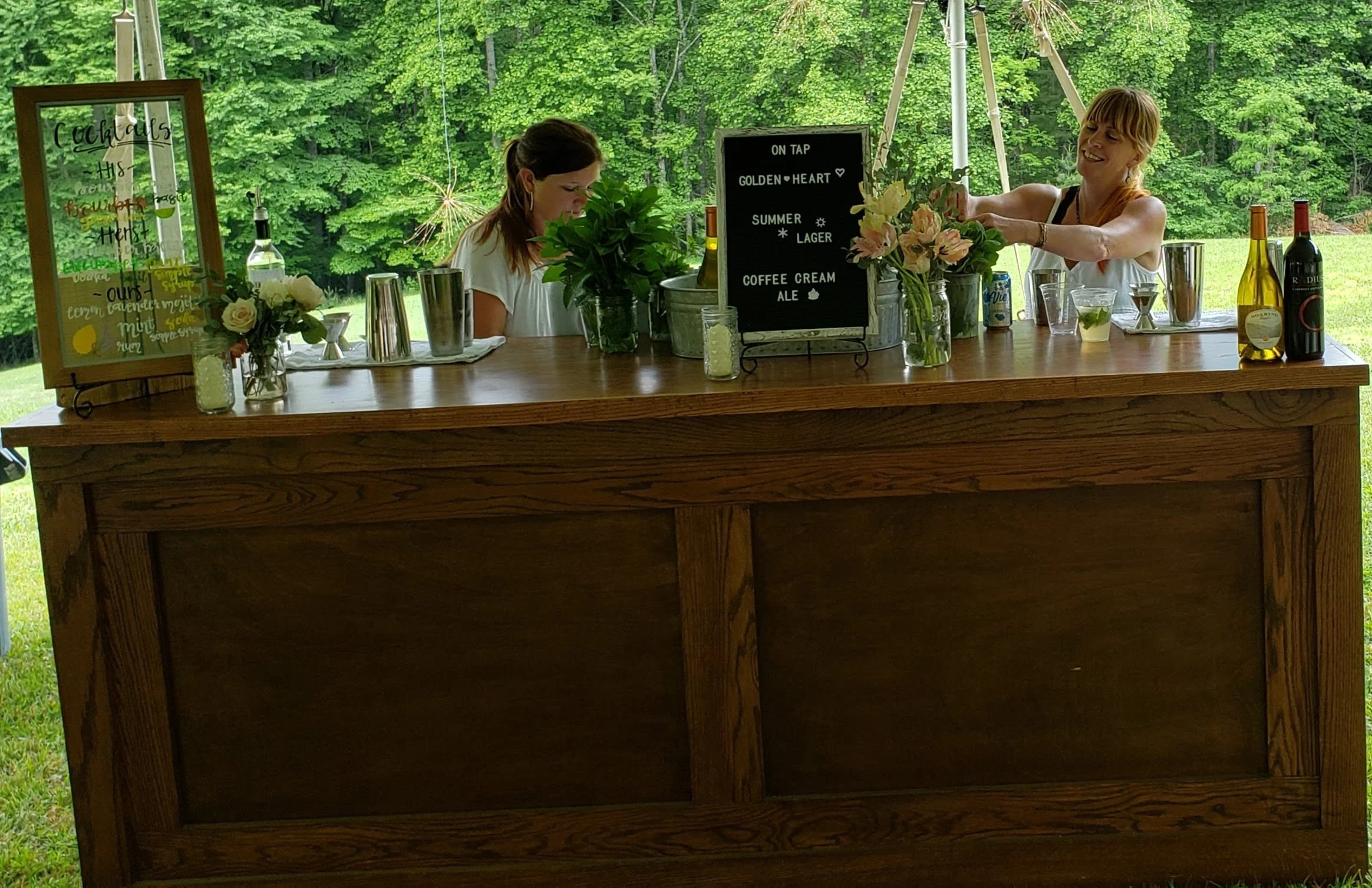 A wooden bar table set up outdoors with flowers, a chalkboard sign, and various bottles and glasses, surrounded by lush green trees.