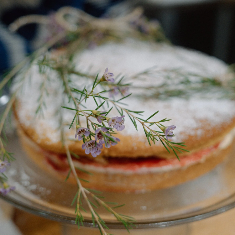 A close-up of a baked cake with powdered sugar and herbs on top, garnished with purple flowering sprigs.