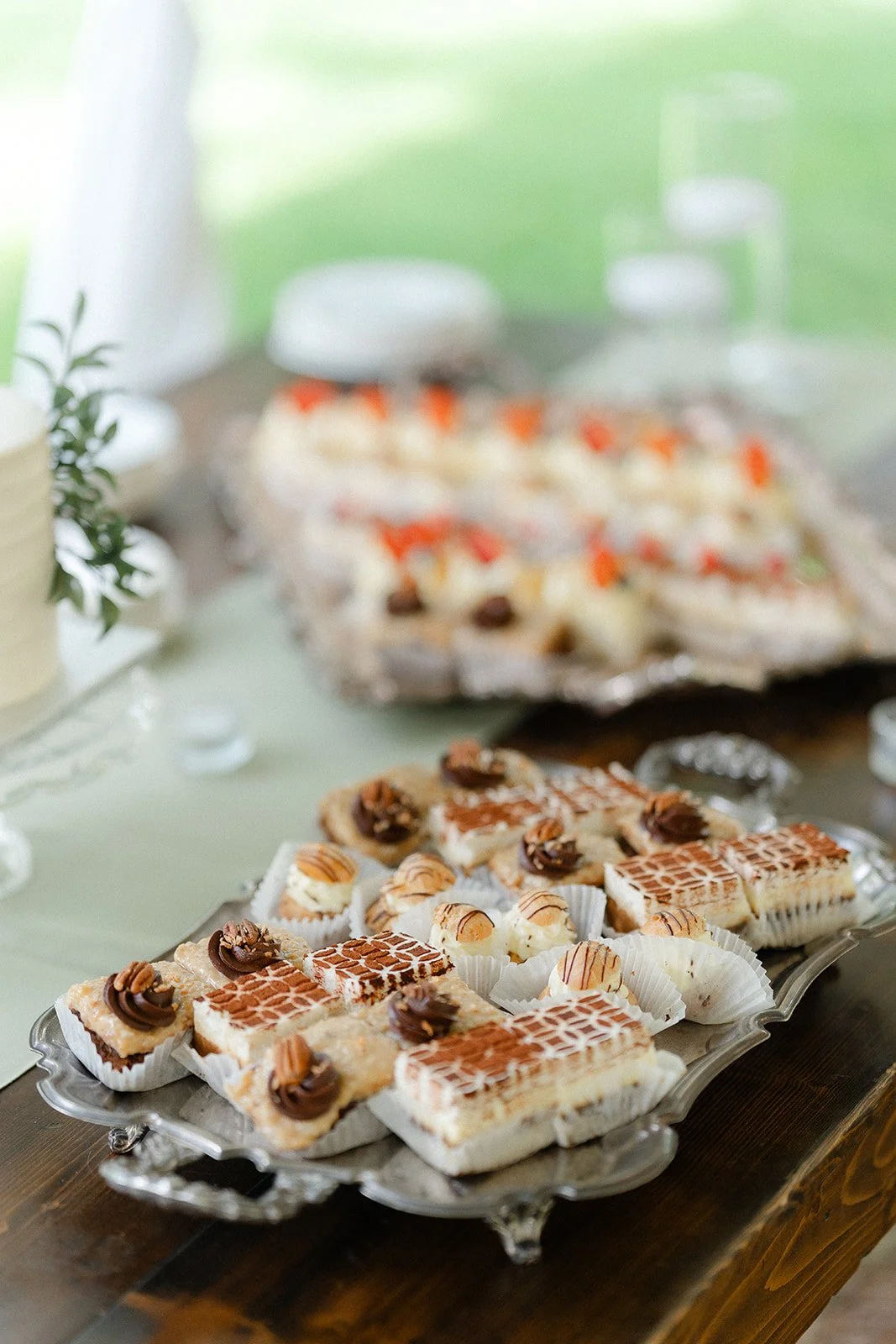 Assorted desserts on a silver platter, including layered cakes, tarts, and pastries topped with nuts and chocolate, on a wooden table.