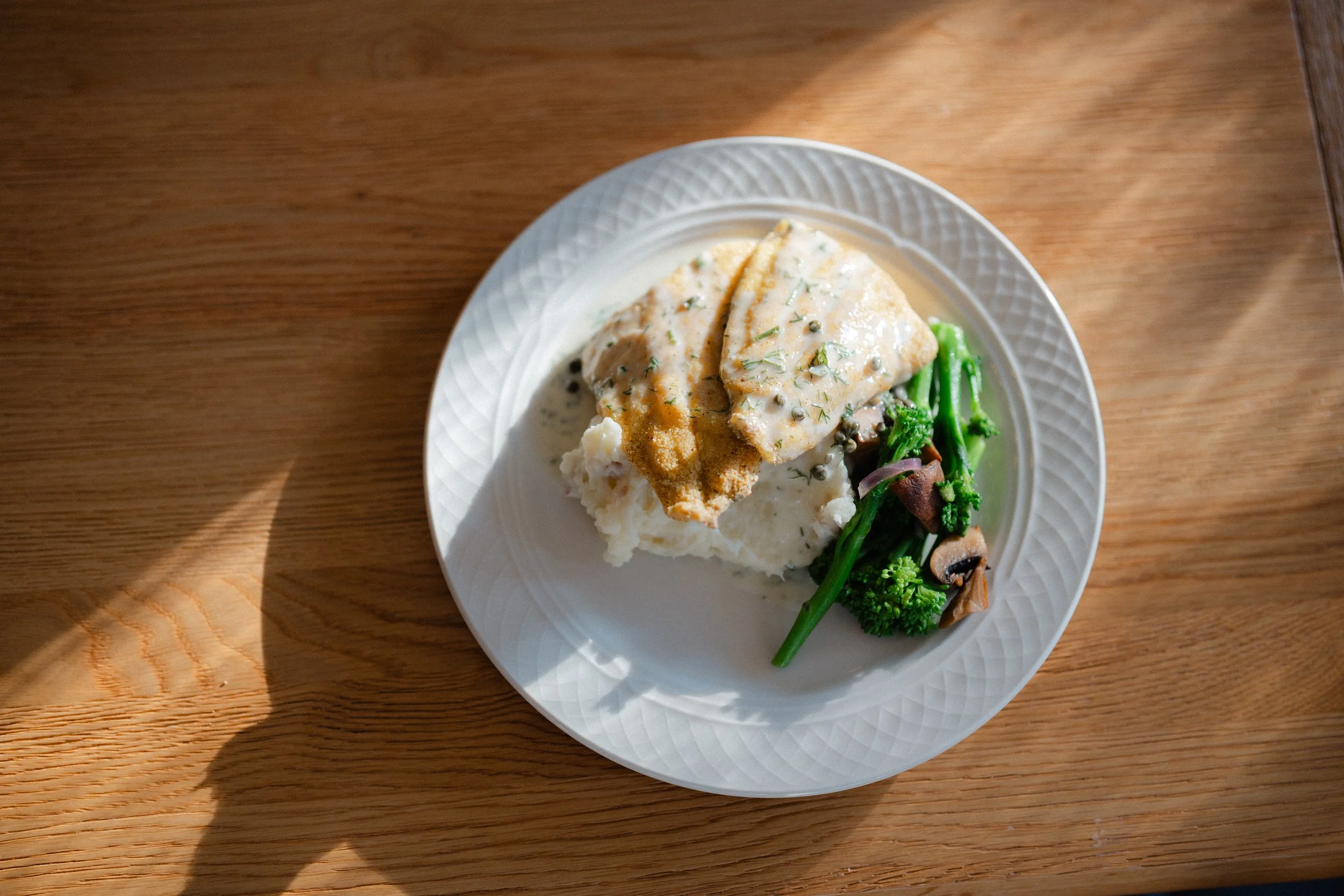 Plate of cooked fish fillet with sauce, mashed potatoes, and steamed broccoli on a wooden table.