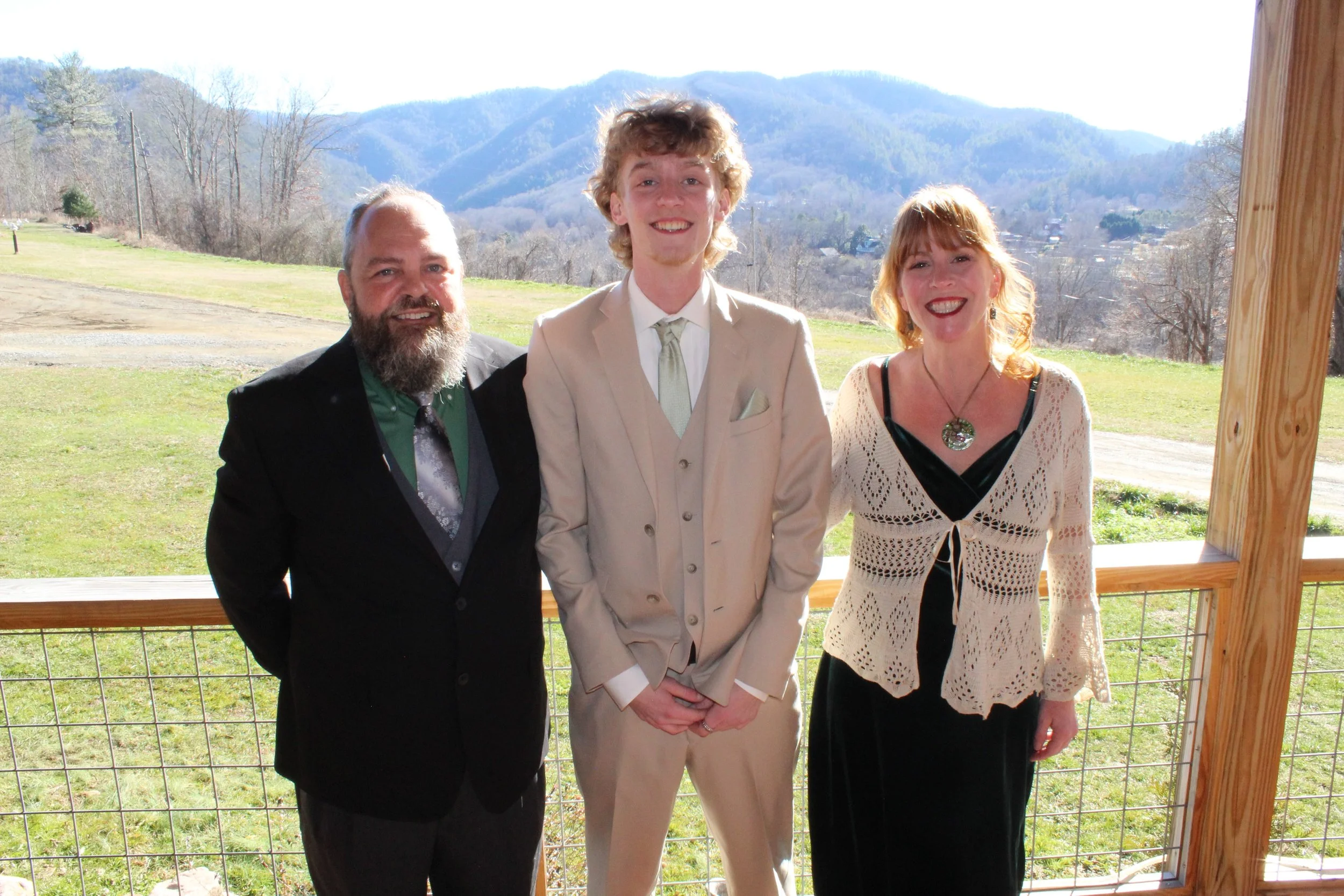 Three people standing outdoors on a balcony with mountains in the background, dressed formally and smiling.
