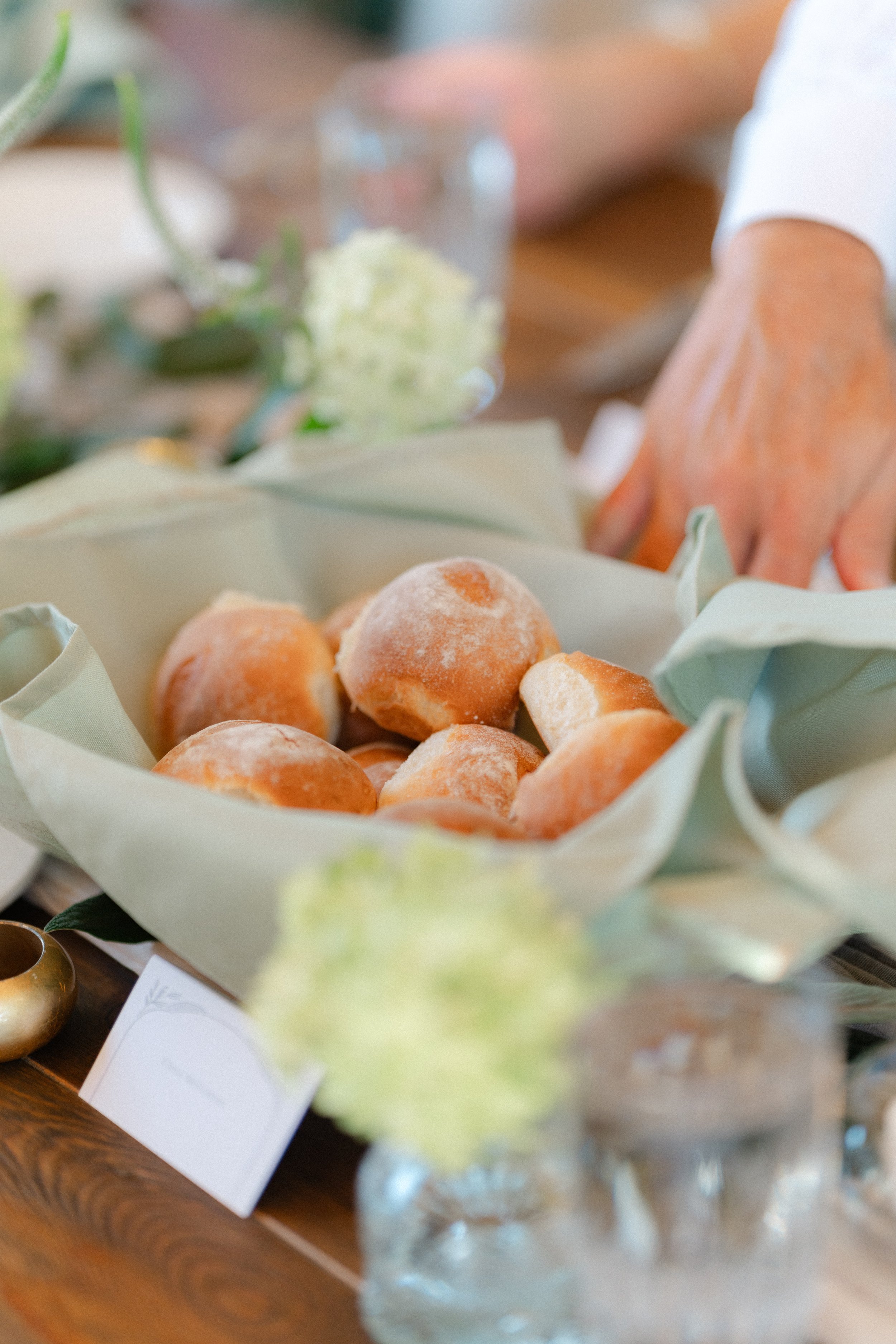 Close-up of a basket of small, round, sugar-coated bread rolls on a wooden table, with a blurred background including a hand and some flowers.