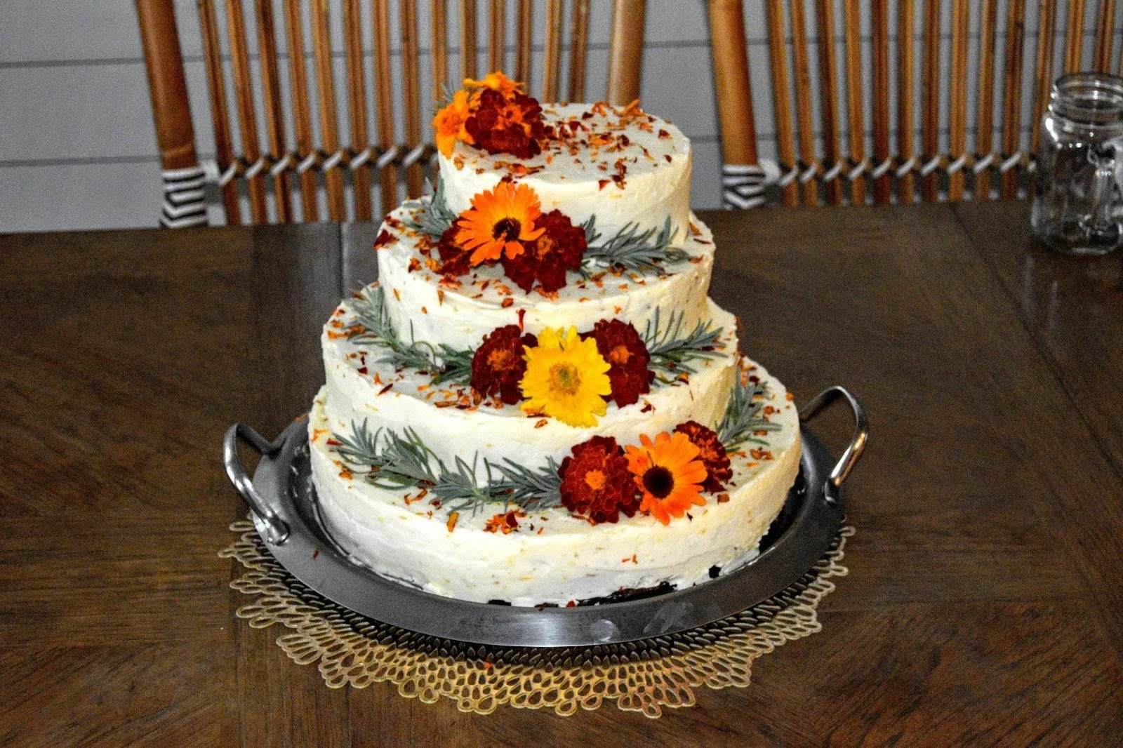 A three-tiered white cake decorated with orange and yellow flowers and green leaves on a metal tray with handles, placed on a wooden table.