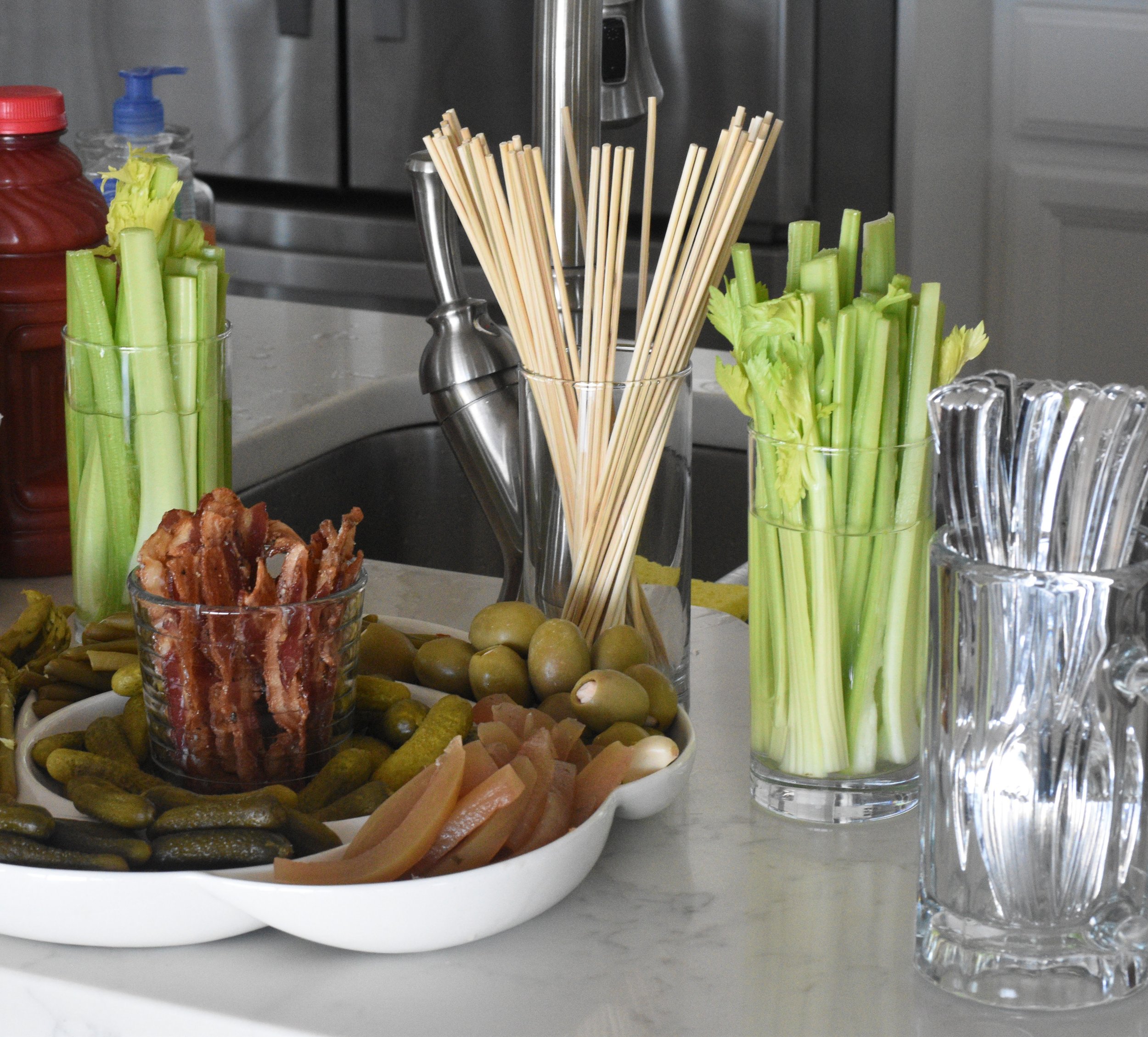 A table with various appetizers including a bowl of pickles, slices of ham, and fried meat, with several glasses filled with celery and pasta, and a plate of olives in the background.