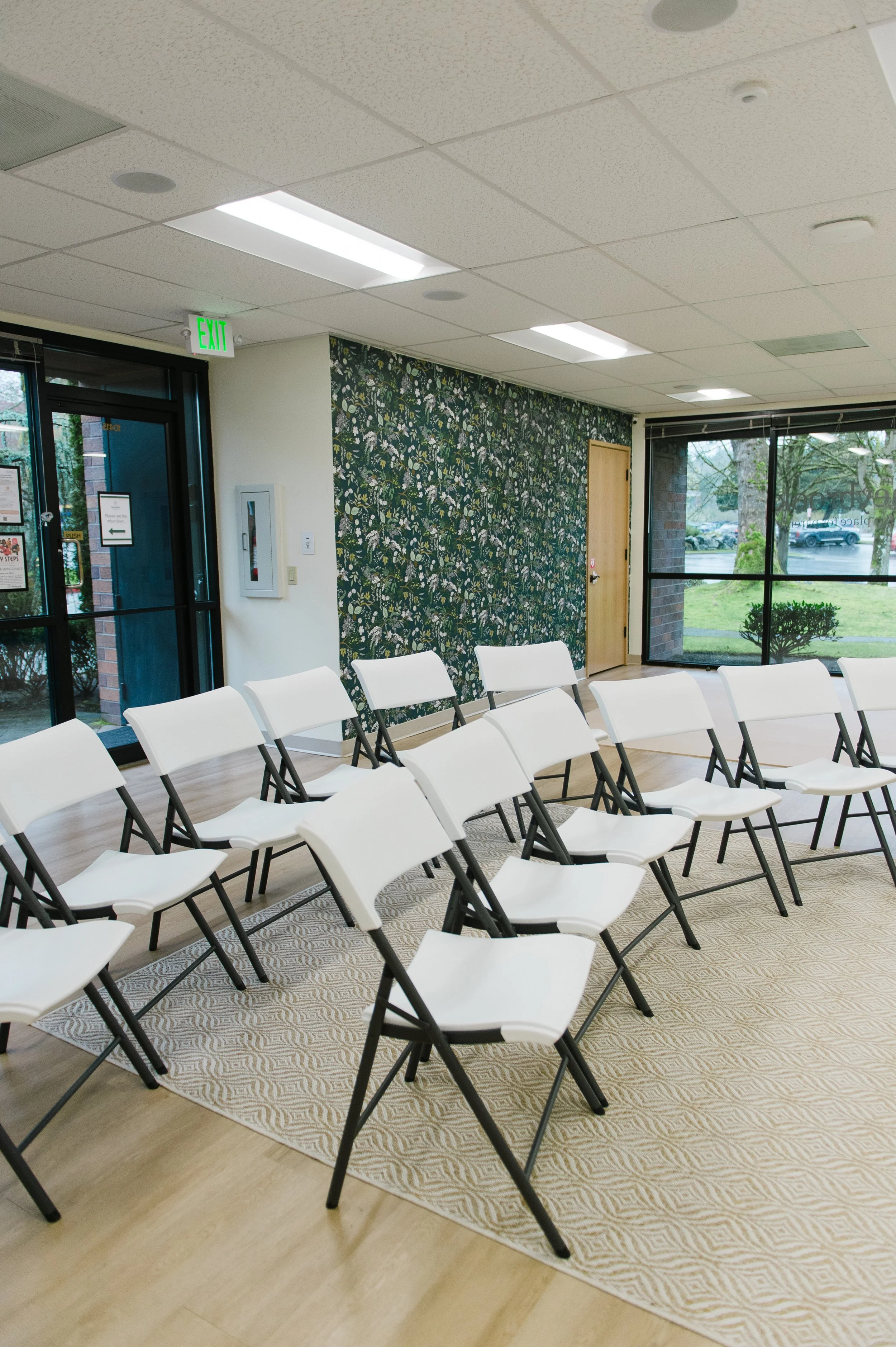 A room set up with white folding chairs arranged in rows on a beige patterned area rug. There are windows with a view of trees and parked cars outside. The room has a floral wallpaper accent wall, a closed wooden door, and fluorescent ceiling lights.