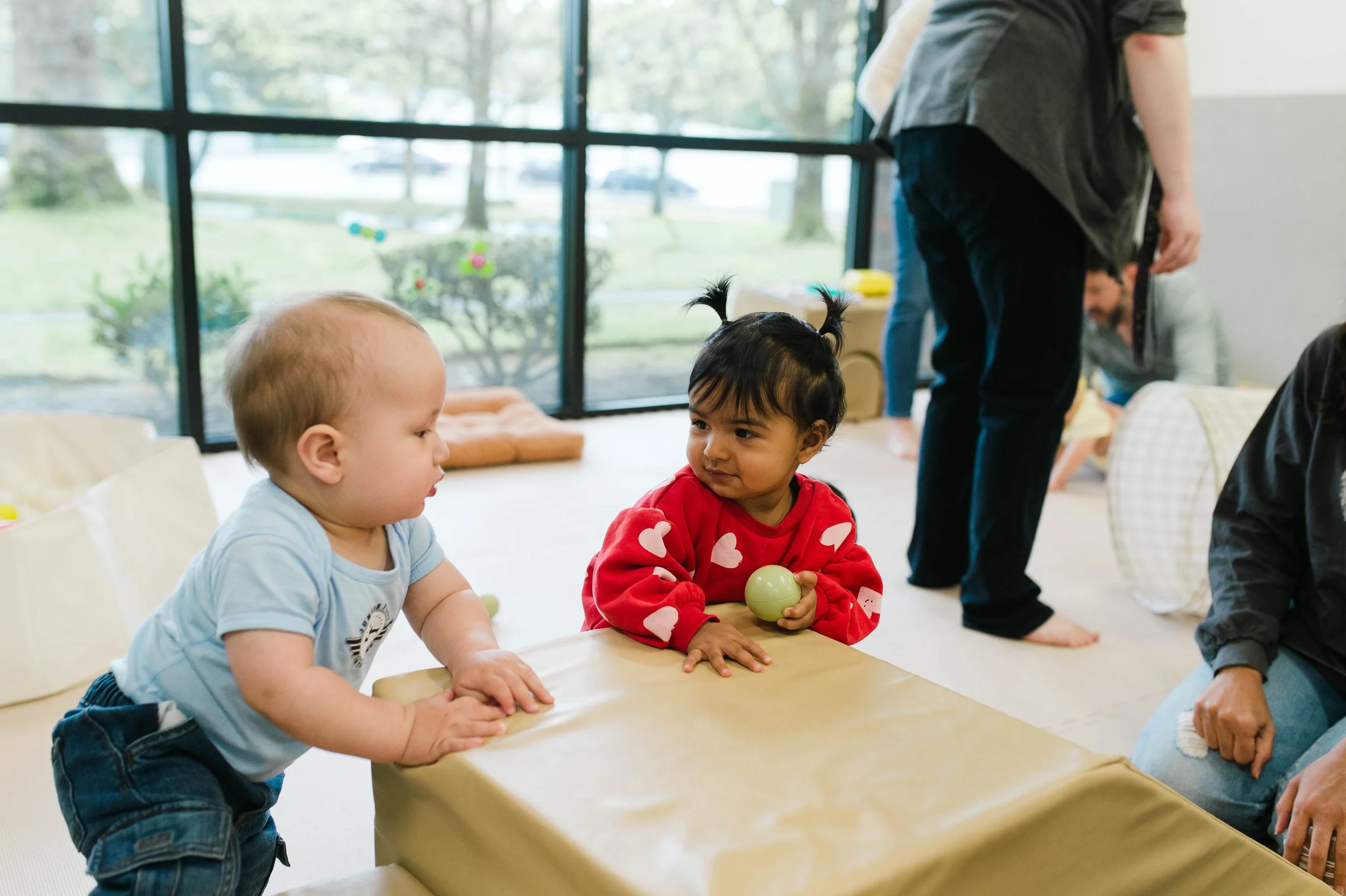 Two young children, a boy and a girl, are sitting at a table. The boy is wearing a light blue shirt and dark jeans, and the girl is wearing a red sweatshirt with white hearts, holding a green ball. They are indoors near large windows, and there are adults in the background.