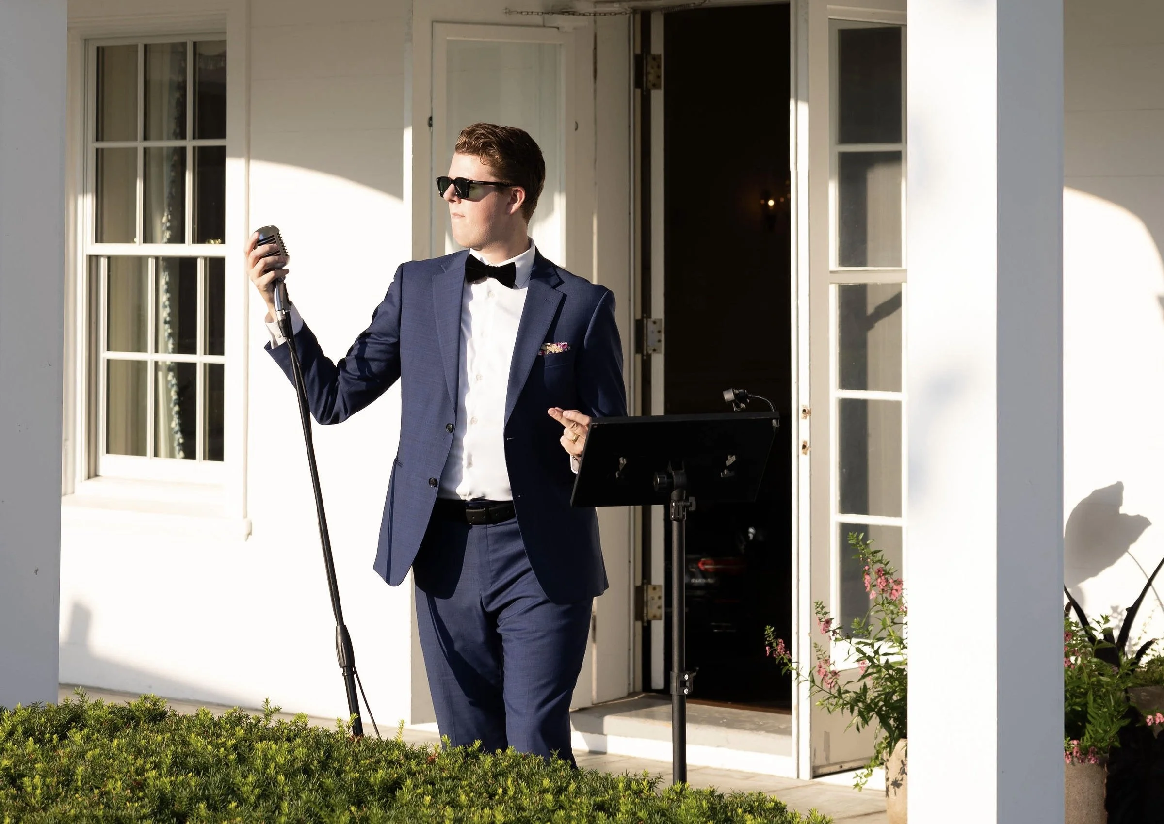 Aaron Davis in a blue suit, white shirt, black bow tie, and sunglasses singing into a vintage microphone outdoors at the Milwaukee Country Club