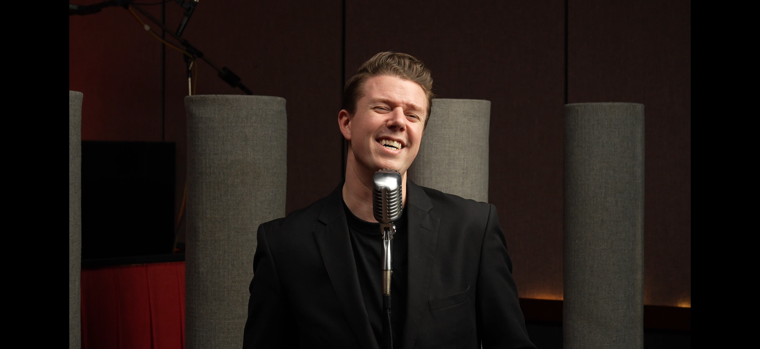 Aaron Davis black suit singing or speaking into a vintage microphone in a recording studio.