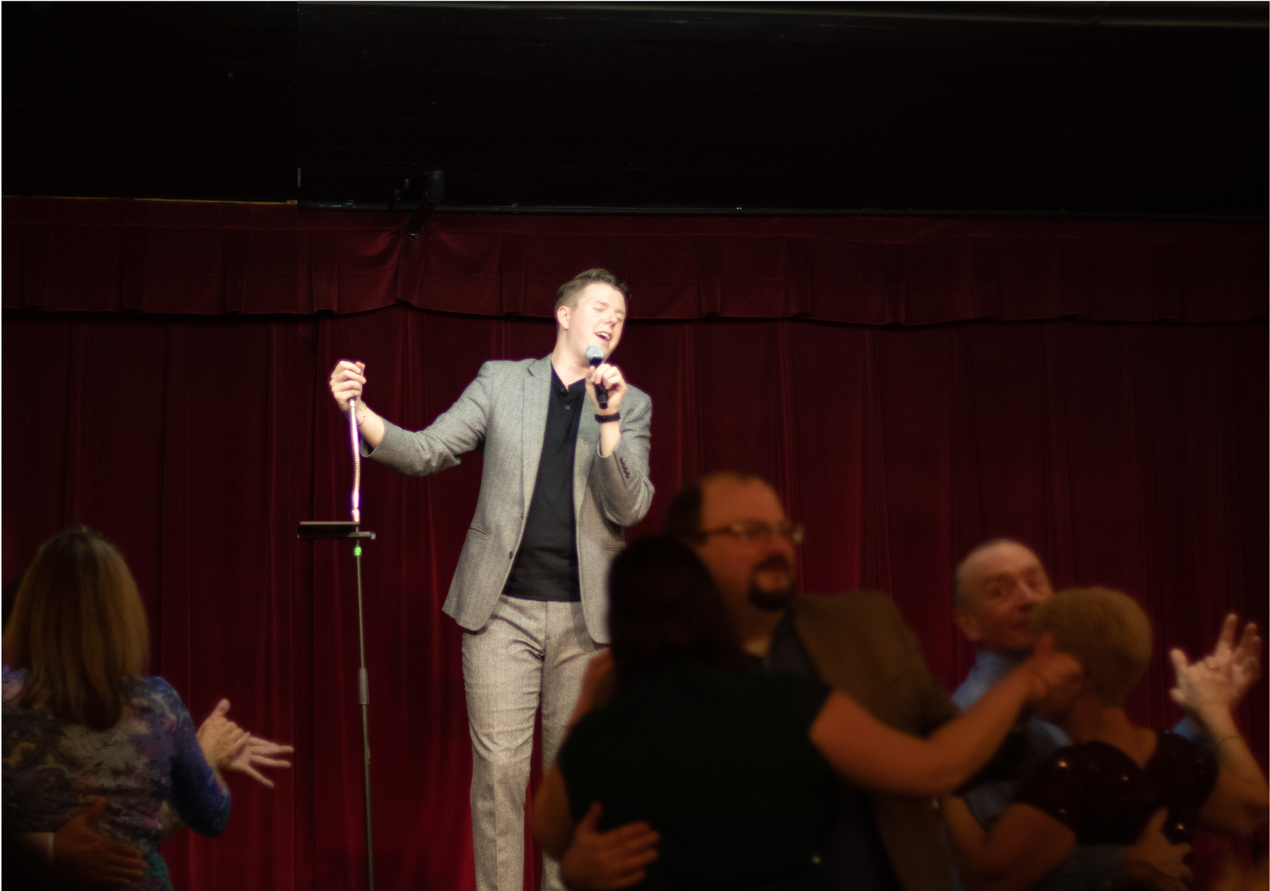 Aaron Davis in a gray suit singing on stage with a microphone, with a red curtain background. Several people are dancing or clapping in front of the stage.