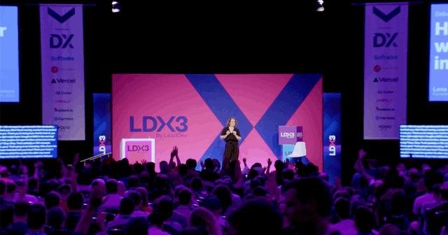 A speaker on stage at a conference with a large audience, screens displaying 'LDX3' and promotional banners over a pink and purple background.